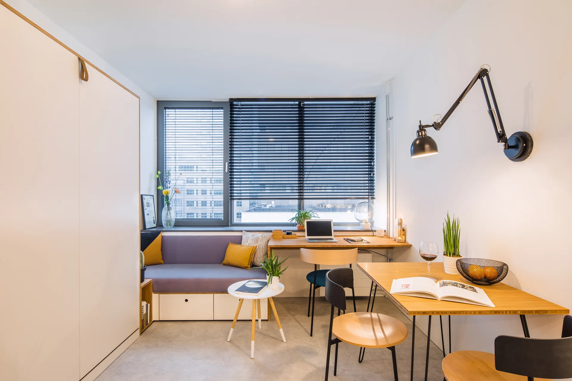 A modern studio apartment features a wooden desk integrated with a purple built-in storage bench under a window with black horizontal blinds.
