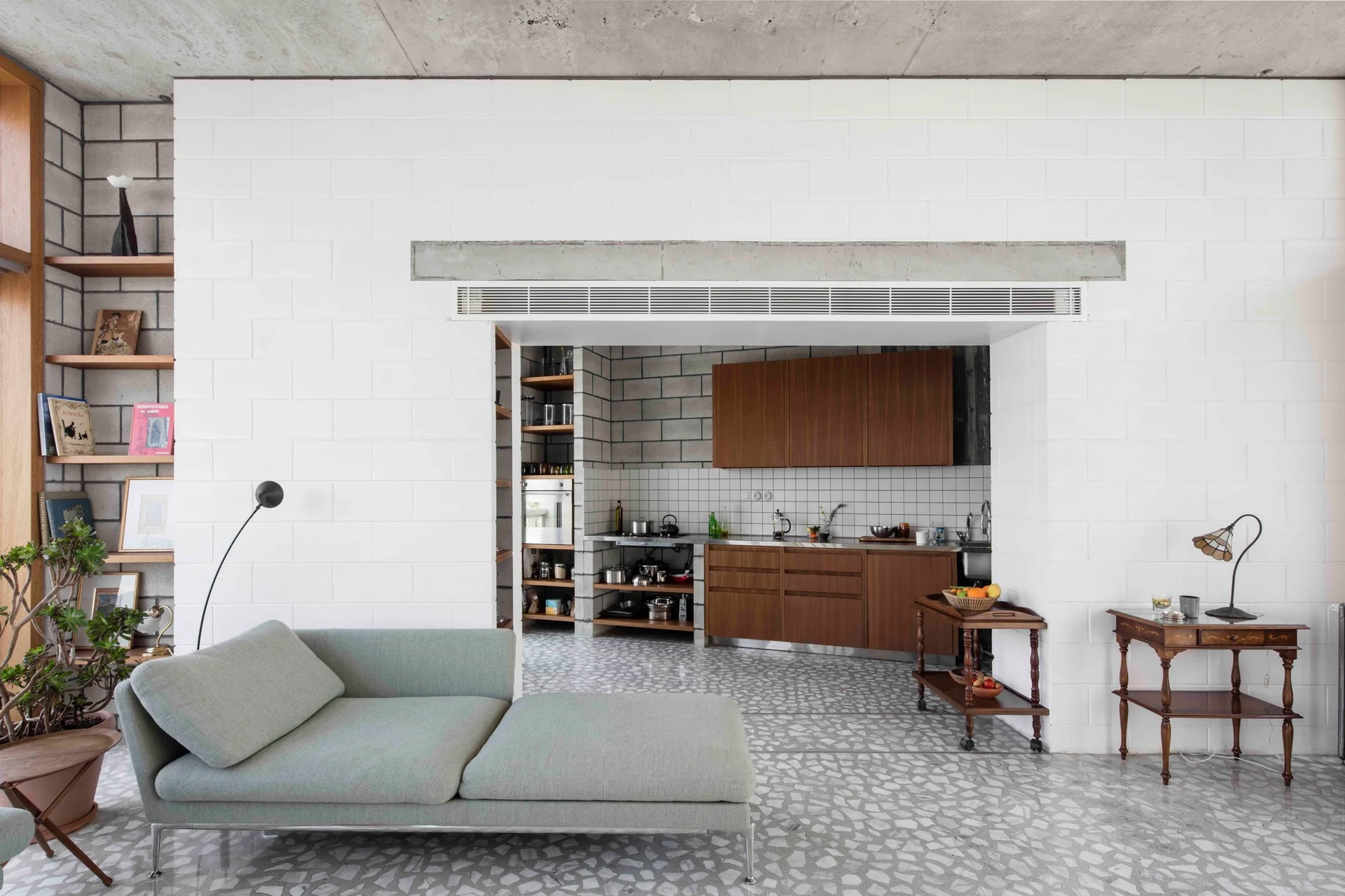 A light gray chaise longue sits in a living room with white cinder block walls, speckled terrazzo flooring, and a view into an adjacent kitchen.