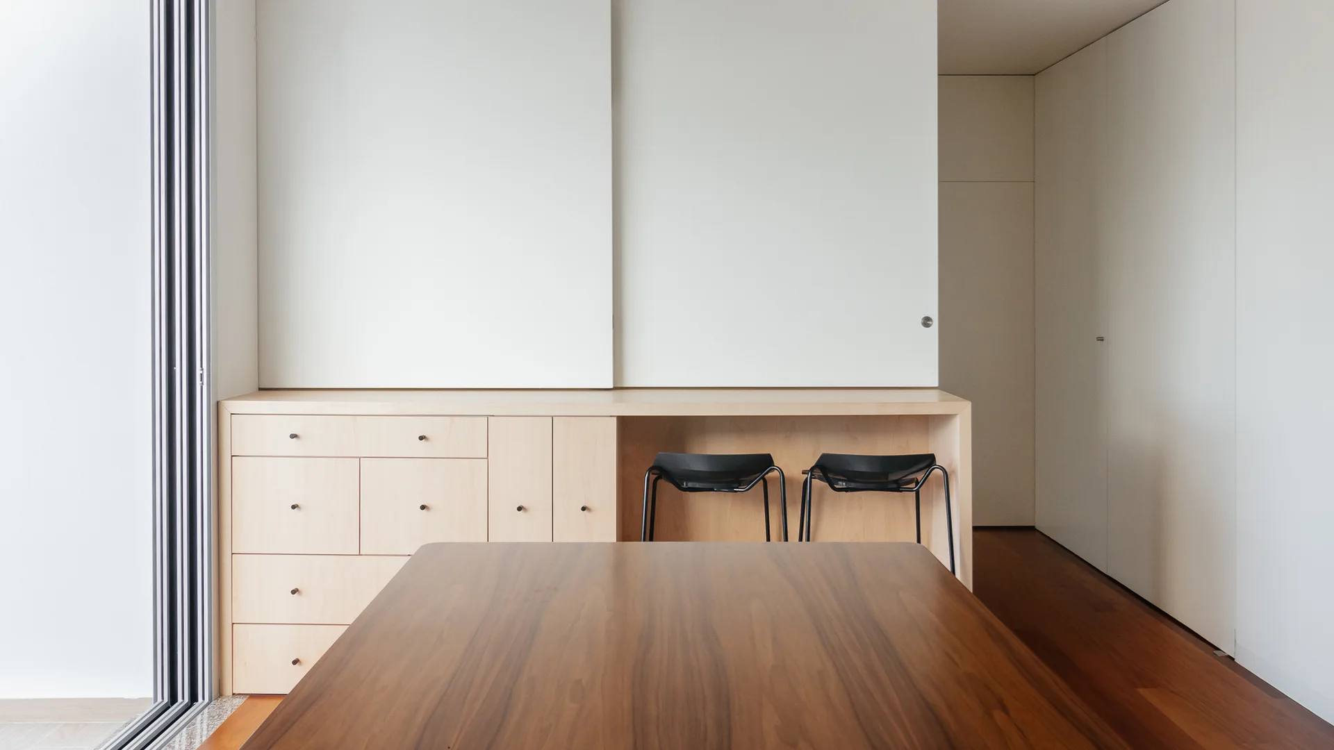 A dark wood table sits in front of light wood cabinetry with a built-in desk niche holding two black metal stools.
