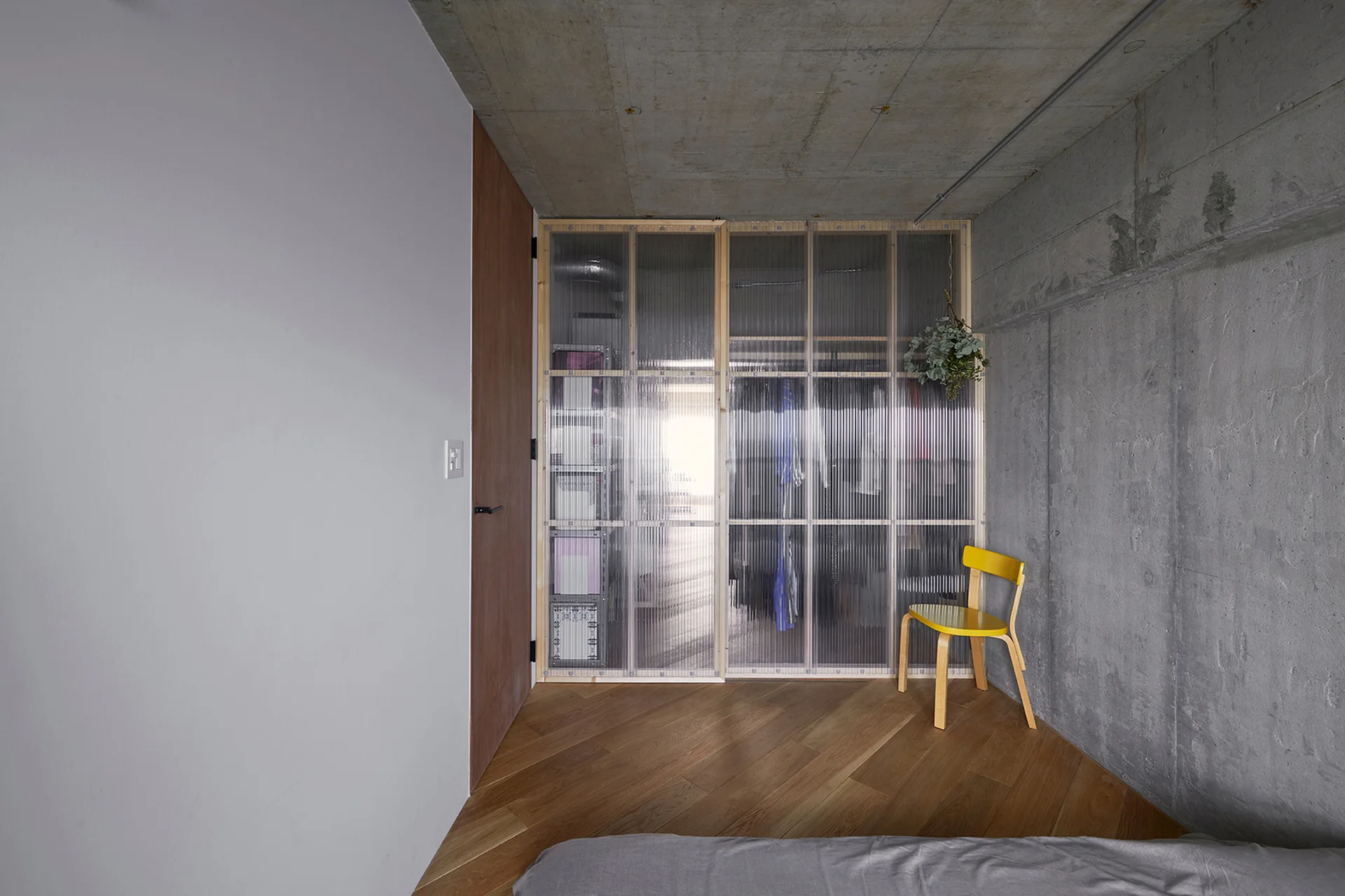 A bedroom featuring an exposed concrete wall, diagonal wood flooring, a bright yellow chair, and a wardrobe with translucent corrugated doors.