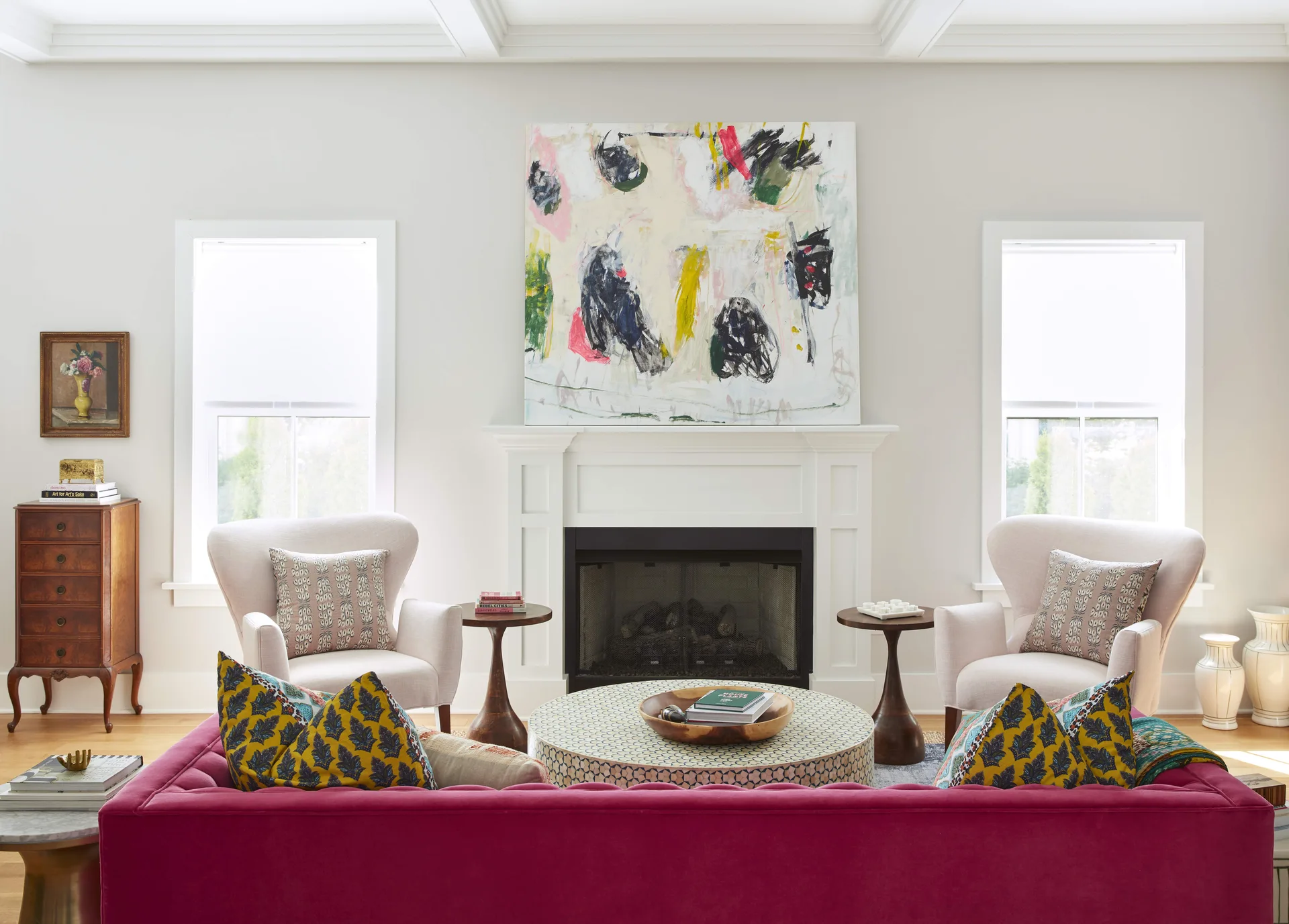 A symmetrical living room features a fuchsia velvet sofa facing a white fireplace mantel topped with a large abstract canvas painting.