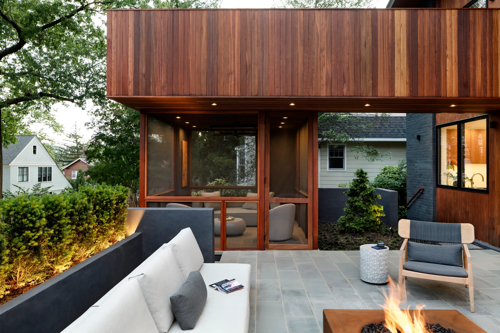 A modern stone patio features a burning fire pit, an outdoor sofa against a grey retaining wall, and an adjacent wood-clad screened-in porch.