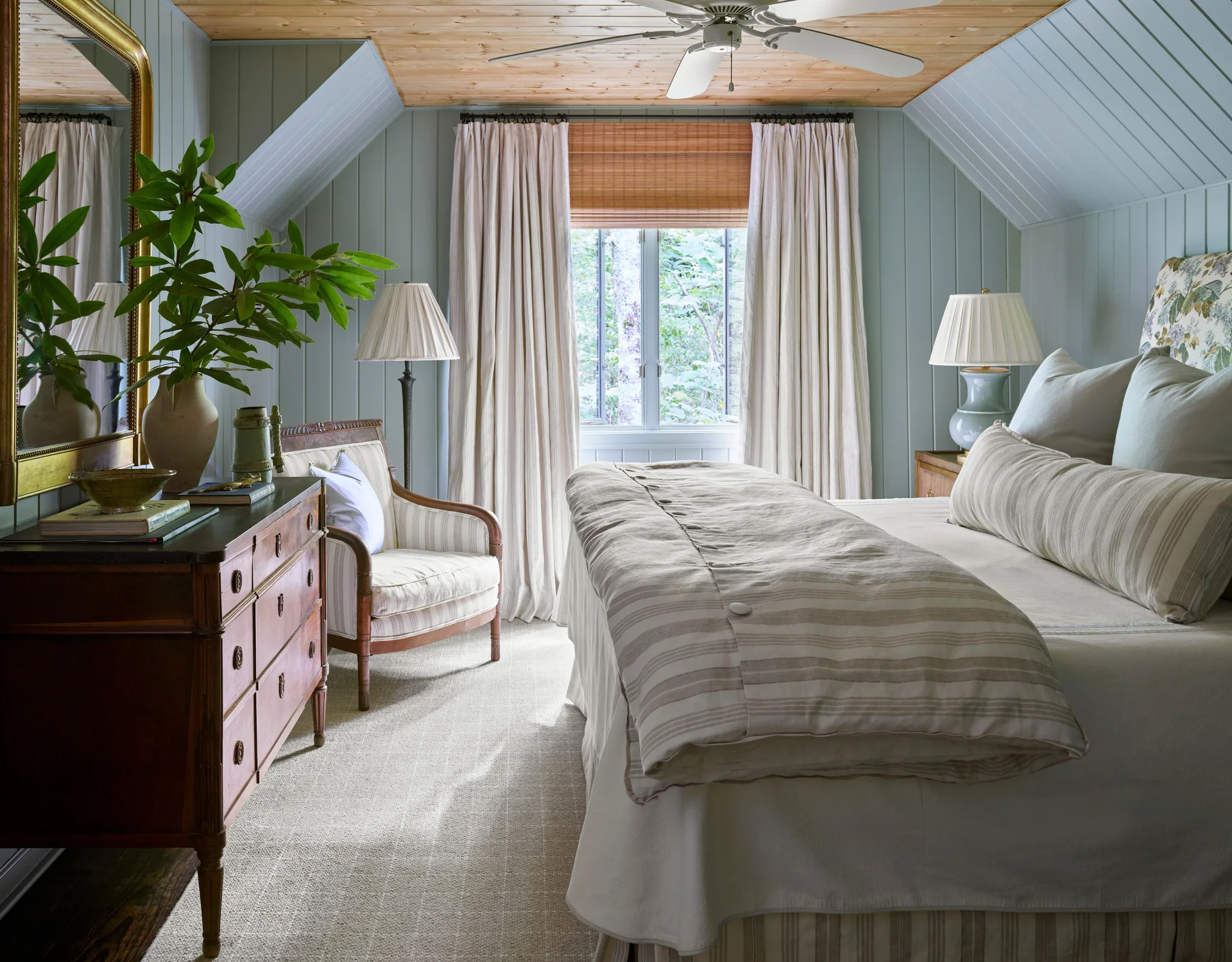 A cozy bedroom with pale blue paneled walls, an exposed wood ceiling, a bed dressed in striped linens, and a vintage wooden dresser.