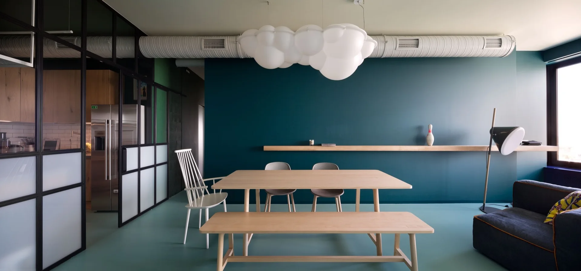 A modern dining room features a light wood table and bench set against a teal wall, with a large cloud-shaped pendant light hanging overhead.