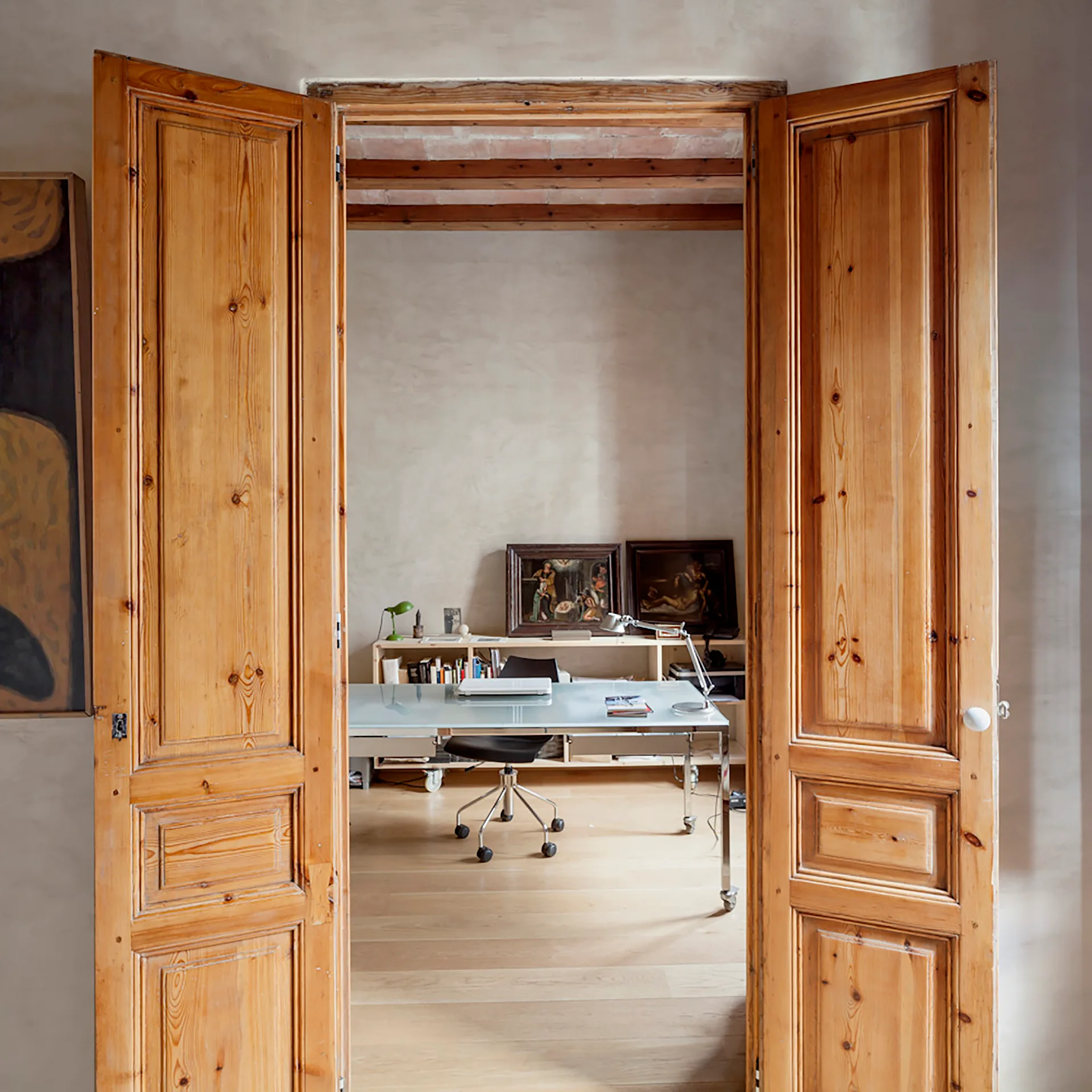 A view through open pine double doors into a minimalist home office with a glass desk, a low wooden bookcase, and exposed ceiling beams.