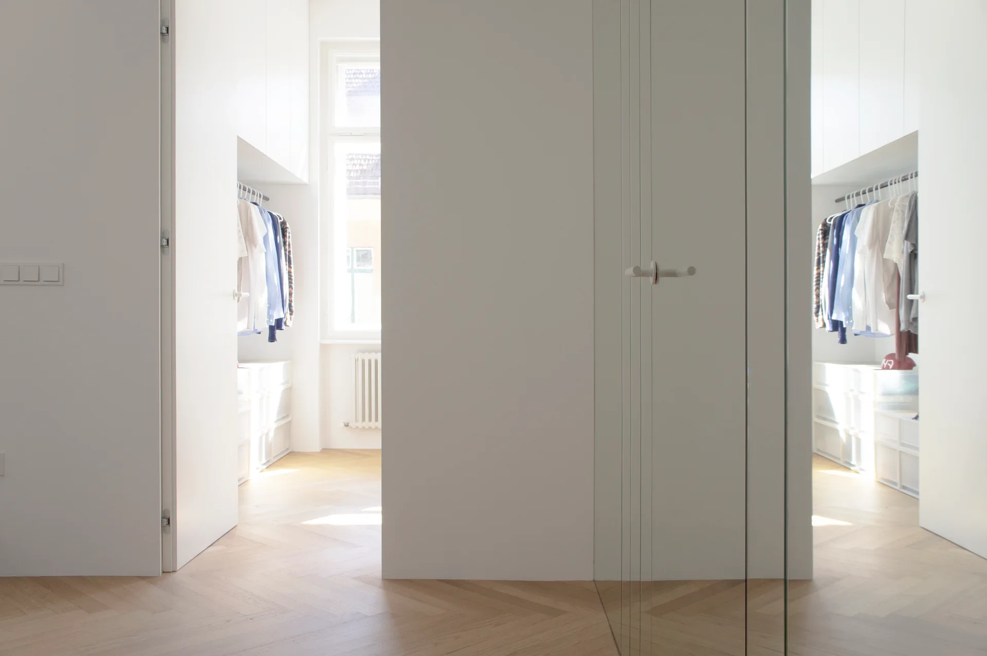 A minimalist white dressing room featuring flush-panel built-in wardrobes, light wood herringbone flooring, and natural light from a window.