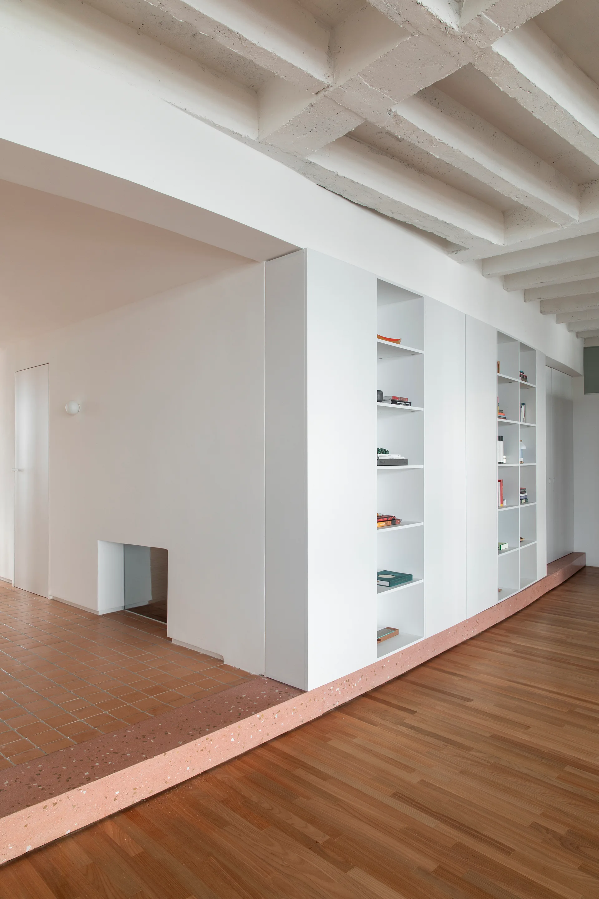 A minimalist room features split wood and tile flooring separated by a pink terrazzo ledge, with custom built-in shelving along the wall.