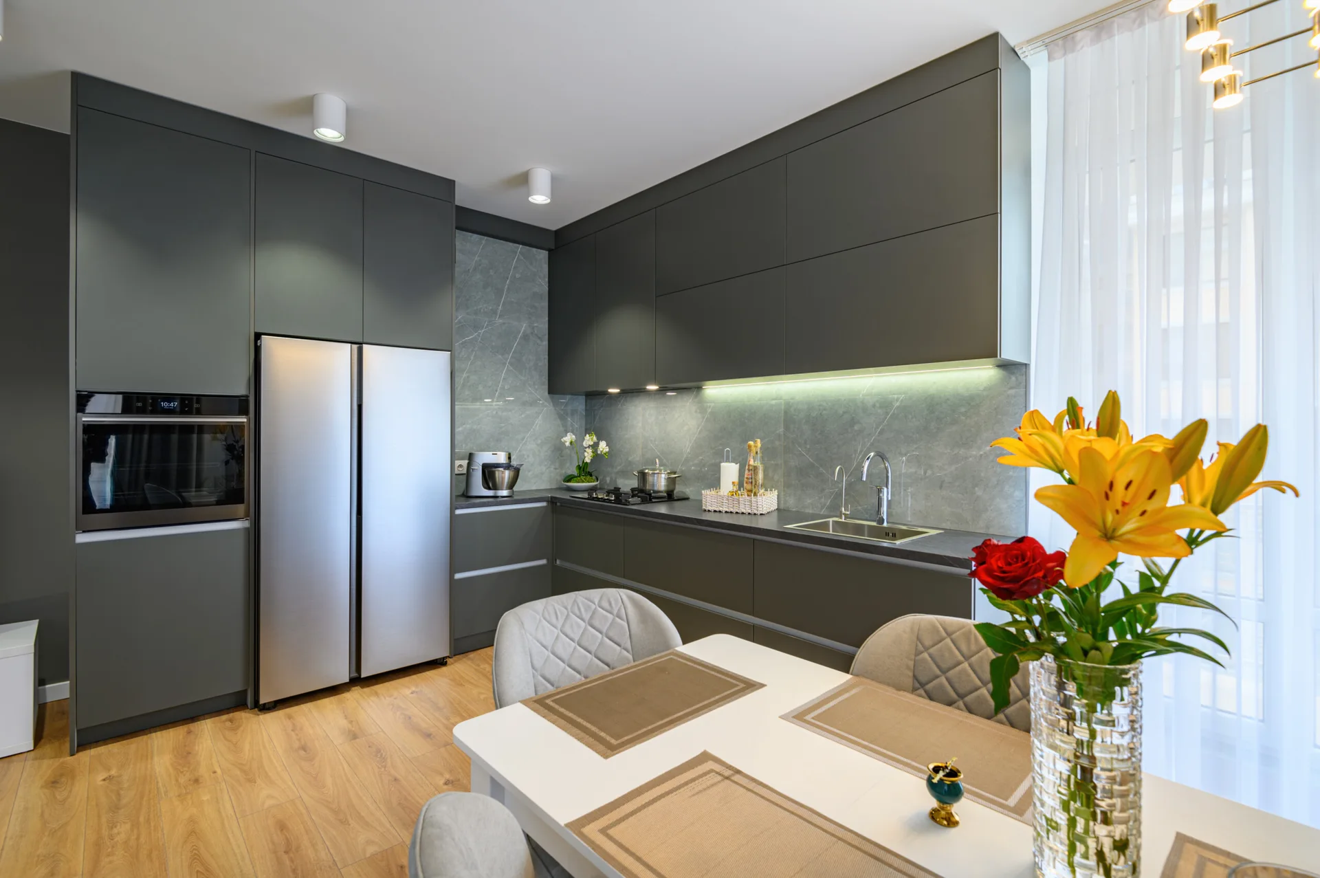 A modern kitchen features matte charcoal grey cabinets, a veined stone backsplash, stainless steel appliances, and a white dining table with quilted chairs.
