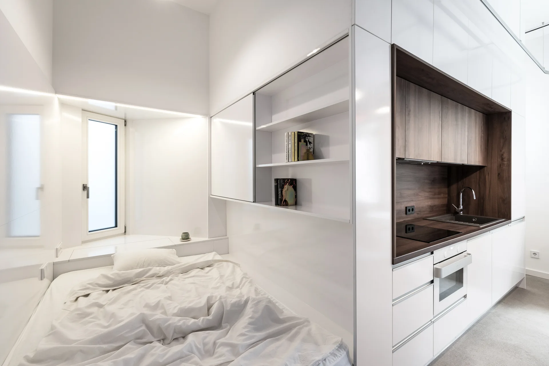 A minimalist studio apartment features a low bed in a built-in nook next to glossy white kitchen cabinets with a dark wood sink area.