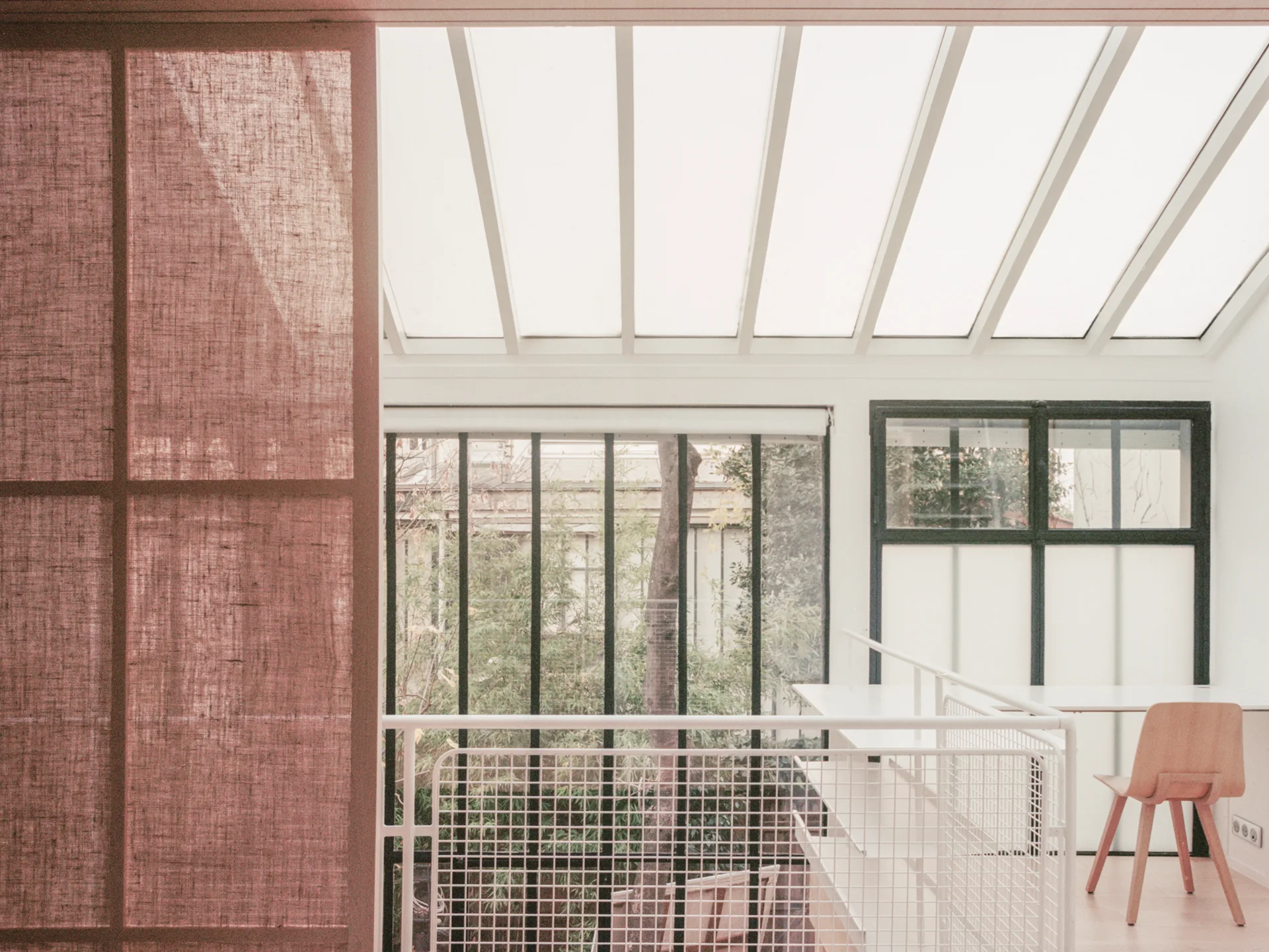 A bright home office features an angled skylight ceiling, a white desk, a wooden chair, and a textured pink screen partition.
