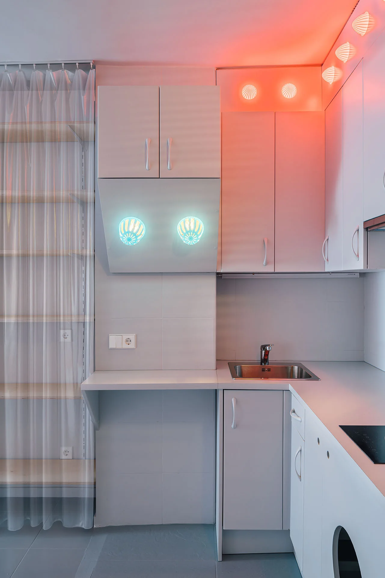A minimalist white kitchen features matte cabinetry, a small steel sink, and shell-shaped wall sconces emitting cyan and orange light.