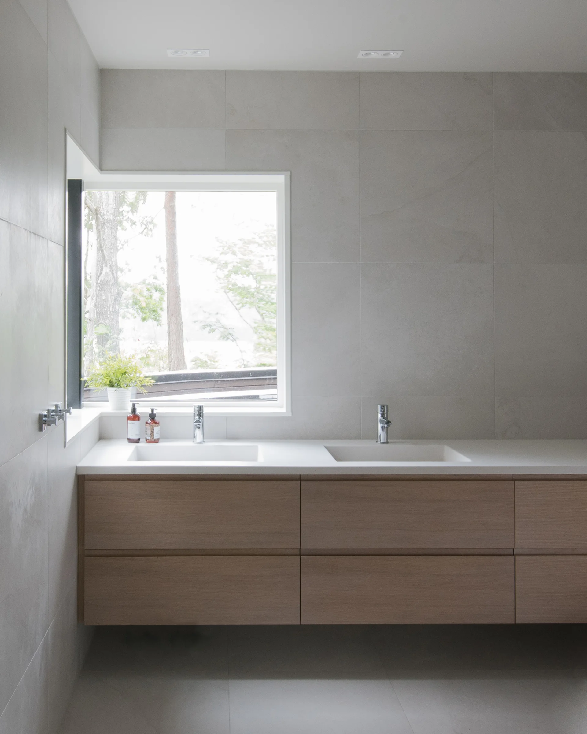 A modern floating wood double vanity stands against a large-format grey tile wall under an integrated corner window showing trees outside.