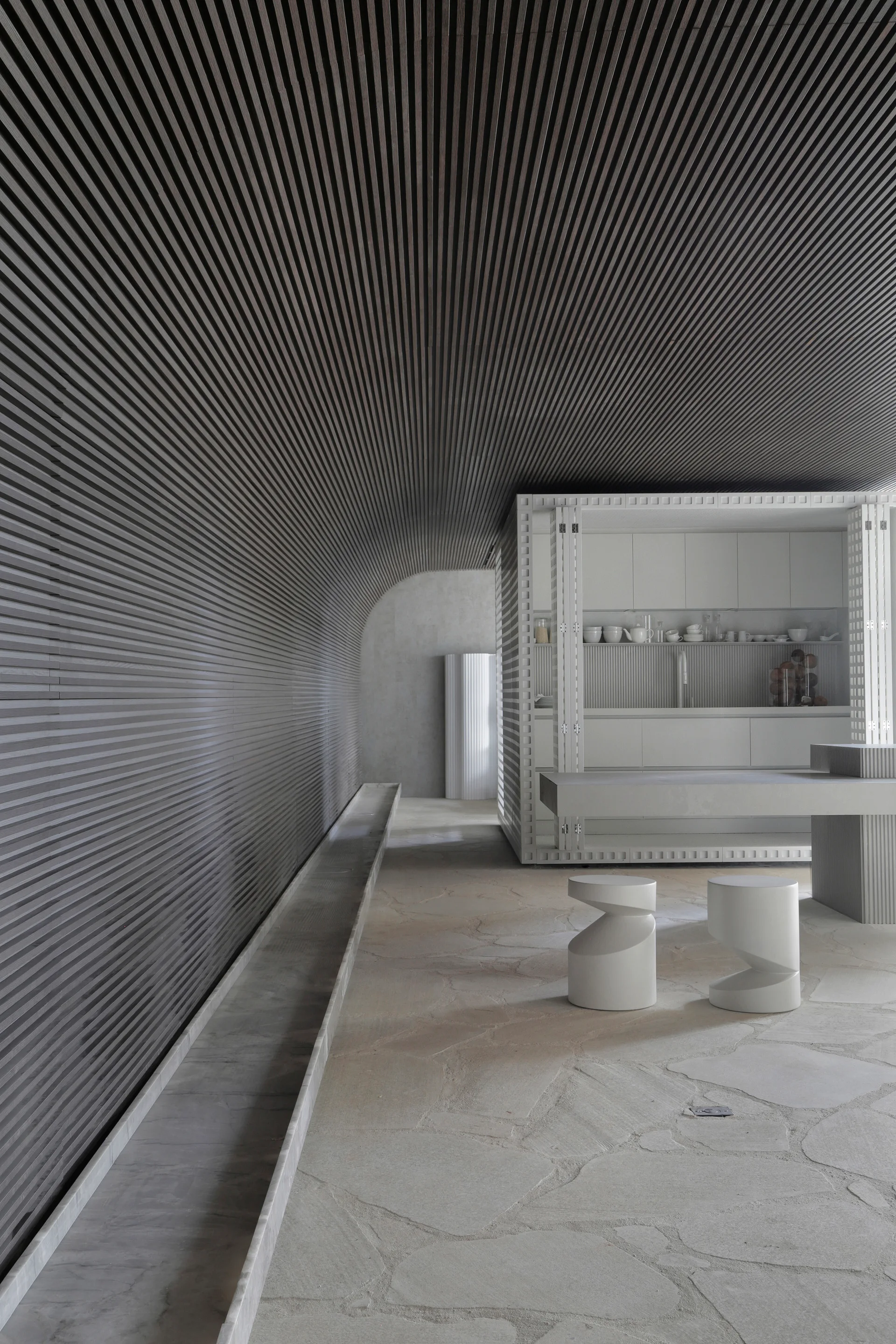 A minimalist kitchen features a curved charcoal slatted wood wall, white cabinetry with folding doors, and irregular flagstone flooring.