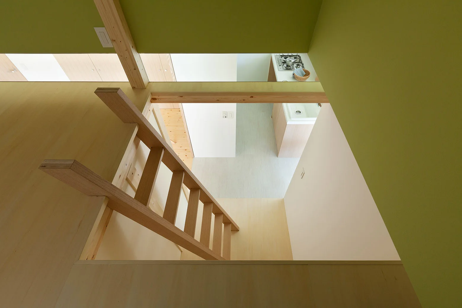 A high-angle view looks down a light wood ladder from a loft space into a minimalist kitchen with white countertops and olive green walls.