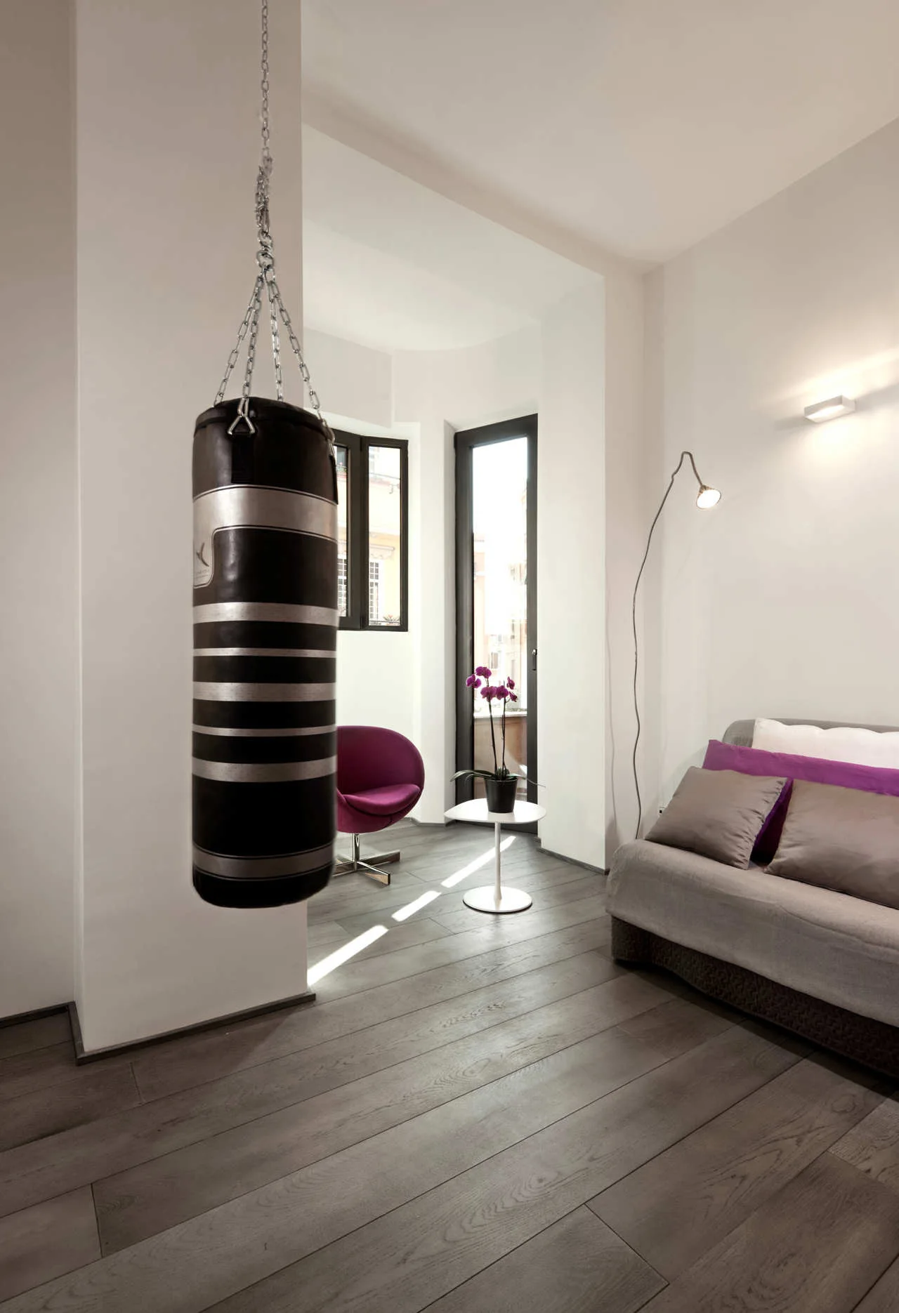 A contemporary room with grey wood floors features a black and silver punching bag hanging in the foreground near a grey sofa with purple pillows.
