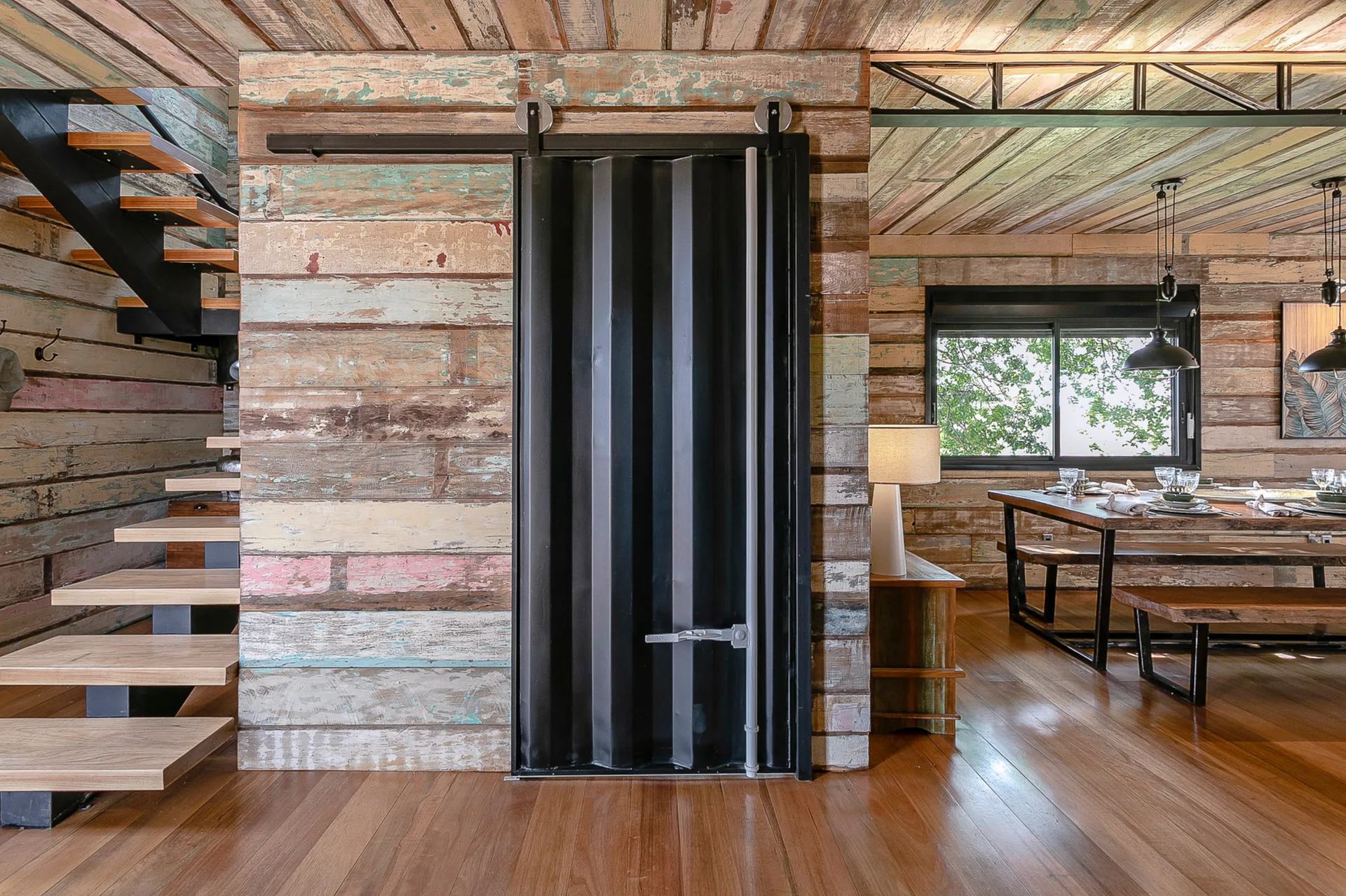A rustic room features multi-colored reclaimed wood walls, floating stairs, and a black corrugated metal sliding door on a track.
