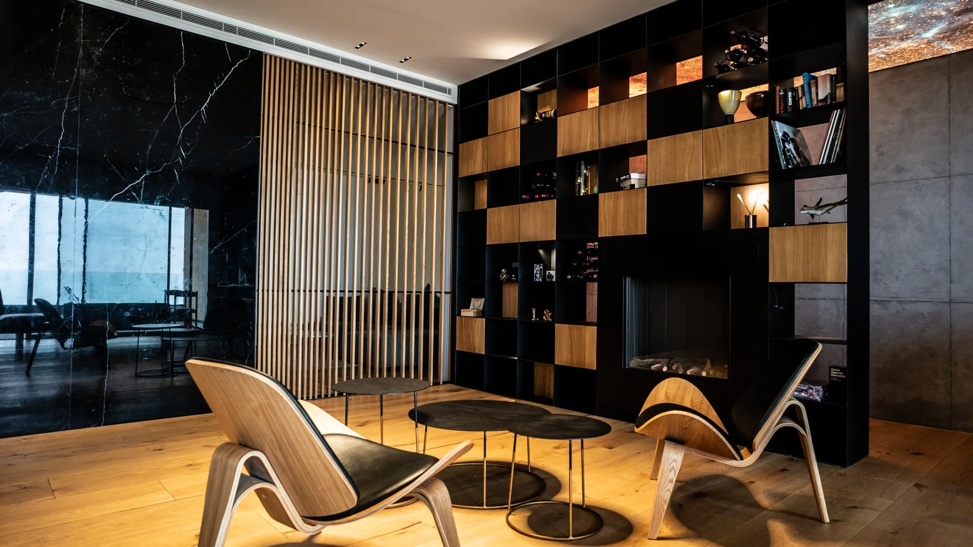 A modern living room features curved wooden armchairs, nesting black metal coffee tables, a wood slat partition, and a large black grid bookshelf.