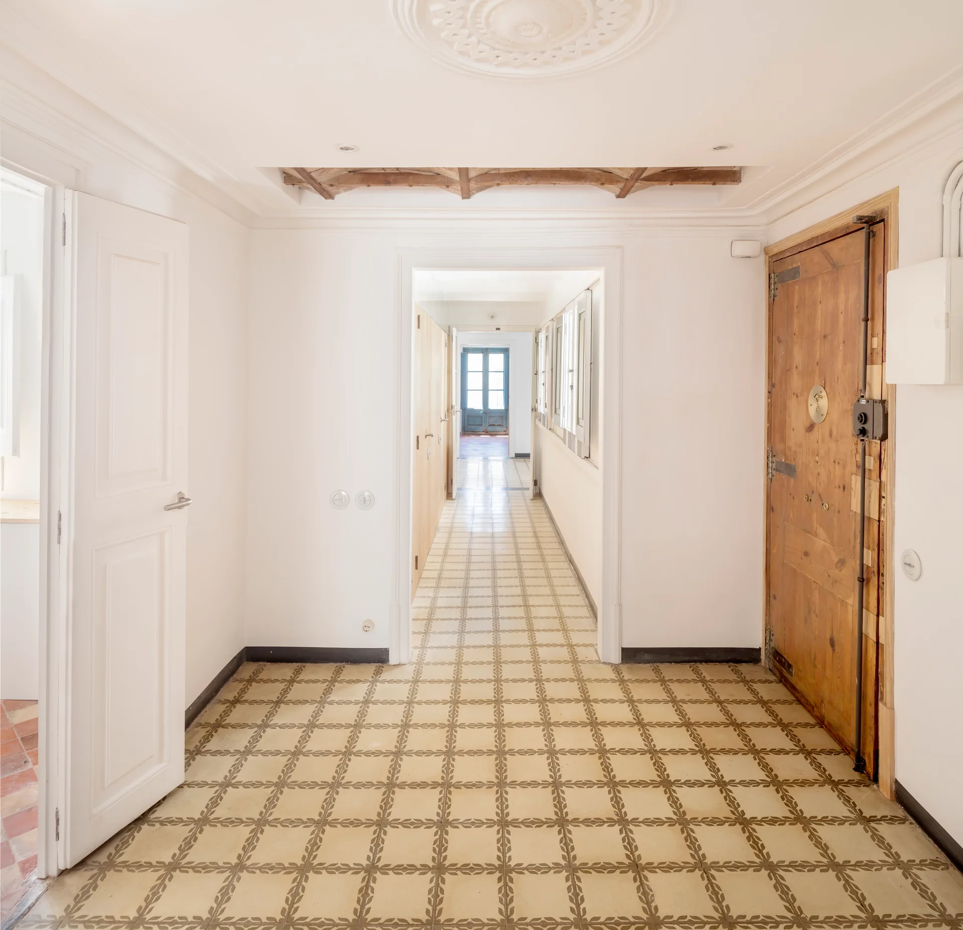 A long white hallway features patterned geometric floor tiles, multiple open doorways, exposed ceiling beams, and a rustic wood door.