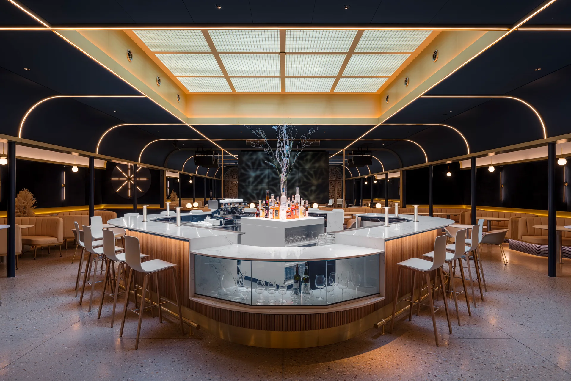 A modern commercial bar featuring a central curved stone-topped counter, white bar stools, and a large illuminated square ceiling grid.
