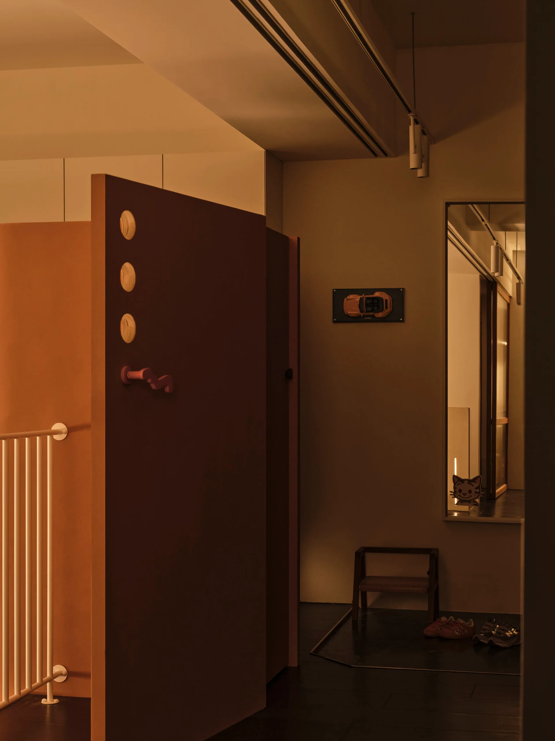 A dimly lit modern hallway features a large maroon partition with circular cutouts beside a recessed entryway with a stool and shoes.