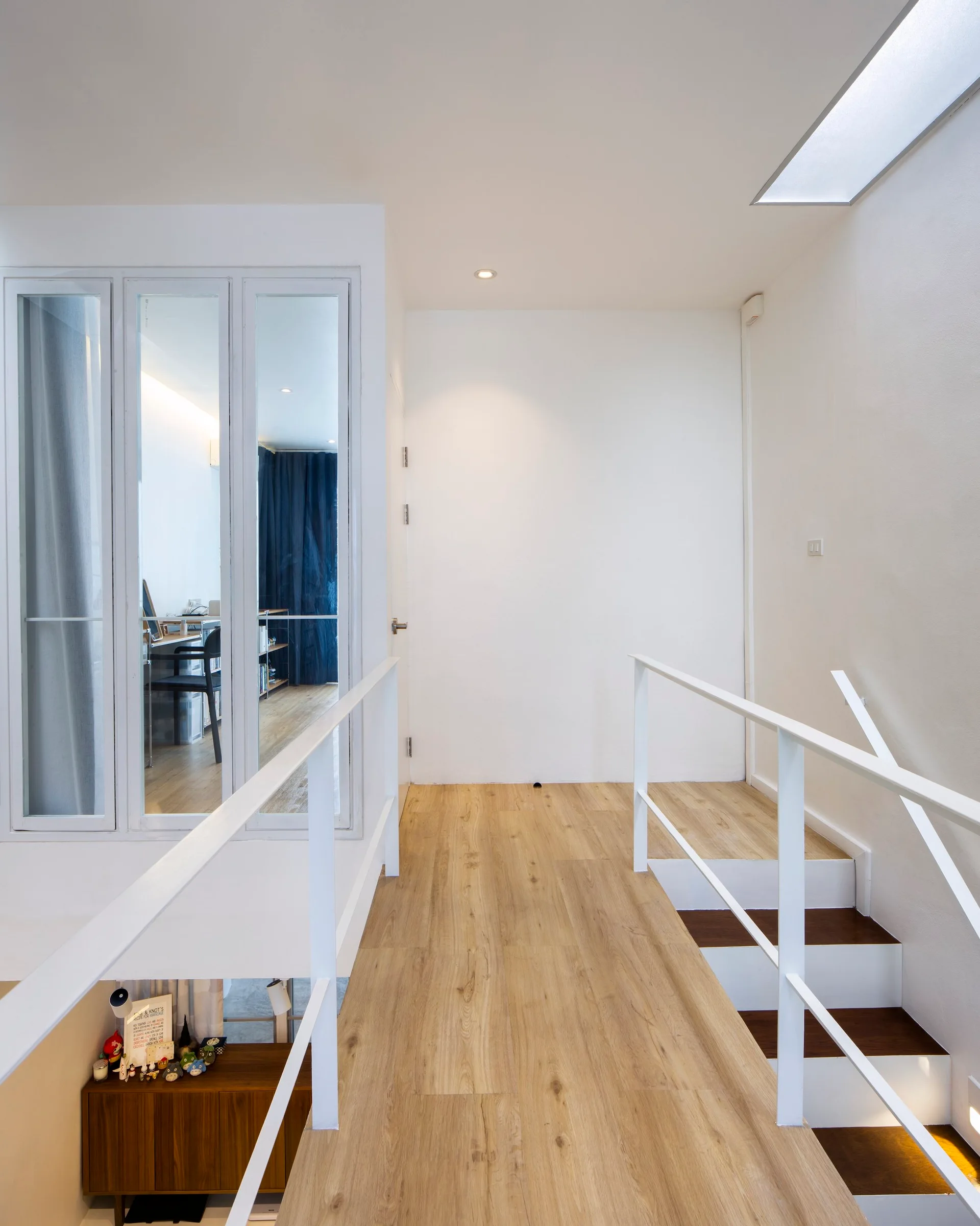 A bright indoor staircase landing with light wood flooring, white minimalist railings, a rectangular skylight, and a glass-enclosed office space.