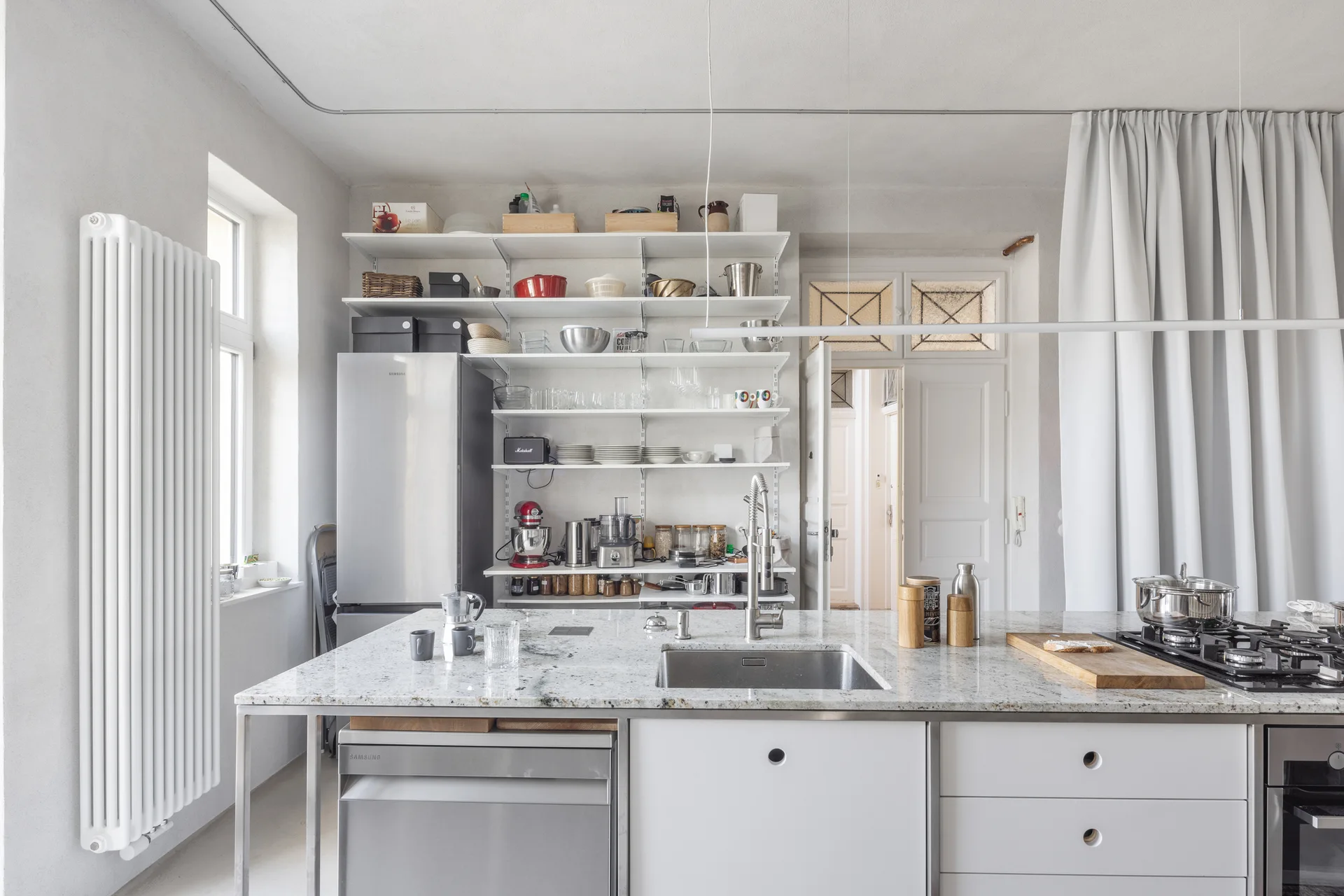 A bright kitchen features a granite-topped island with a sink, stainless steel appliances, and wall-mounted white open shelving holding kitchenware.