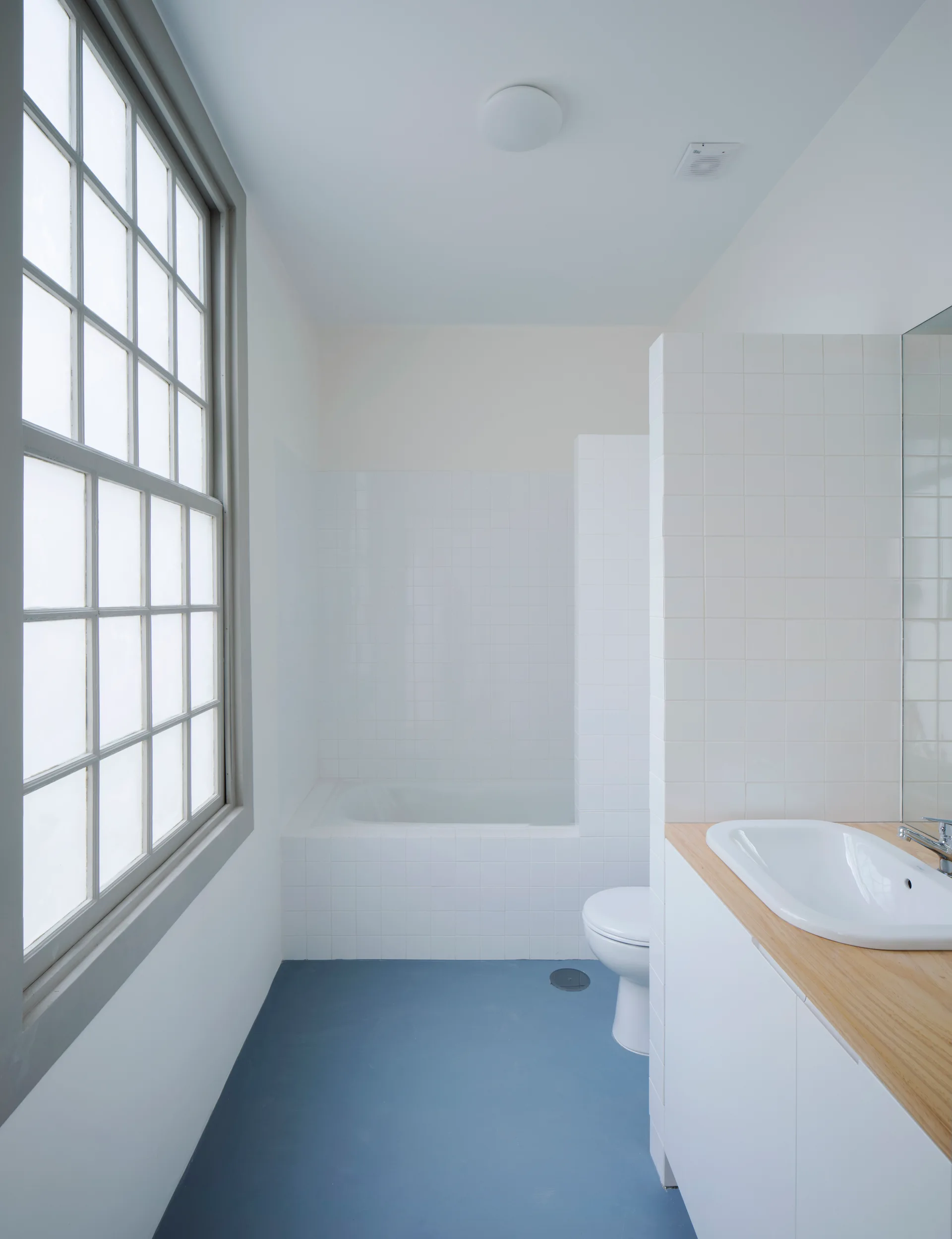 A minimalist bathroom featuring a light blue floor, white tiled walls, a wood-topped vanity with a sink, and a large multi-pane window.