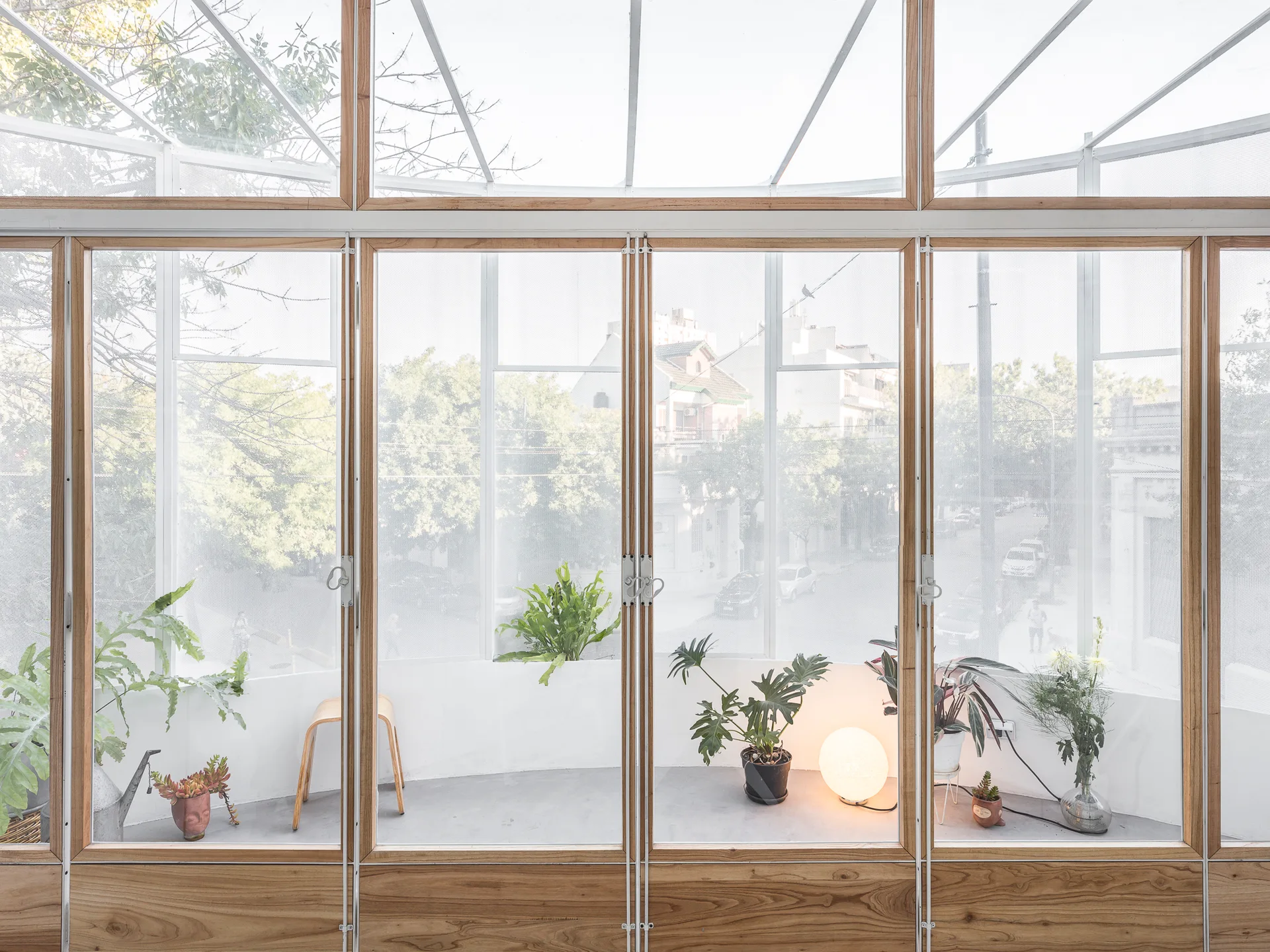 A bright sunroom features large wood-framed glass panels with white mesh screens, potted plants, and a glowing globe lamp on a grey floor.