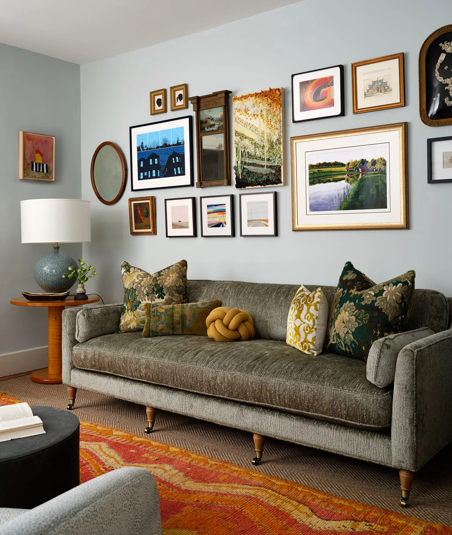 A grey-green sofa rests beneath a dense gallery wall of framed art prints on a light blue wall next to a wooden side table.