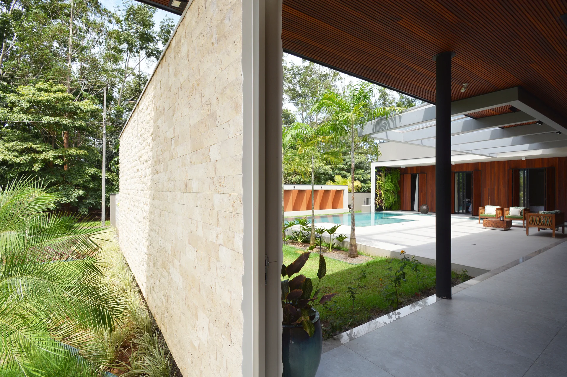 A modern covered patio with a textured stone wall, a dark wood-slatted ceiling, and a concrete tiled floor leading to an outdoor swimming pool.