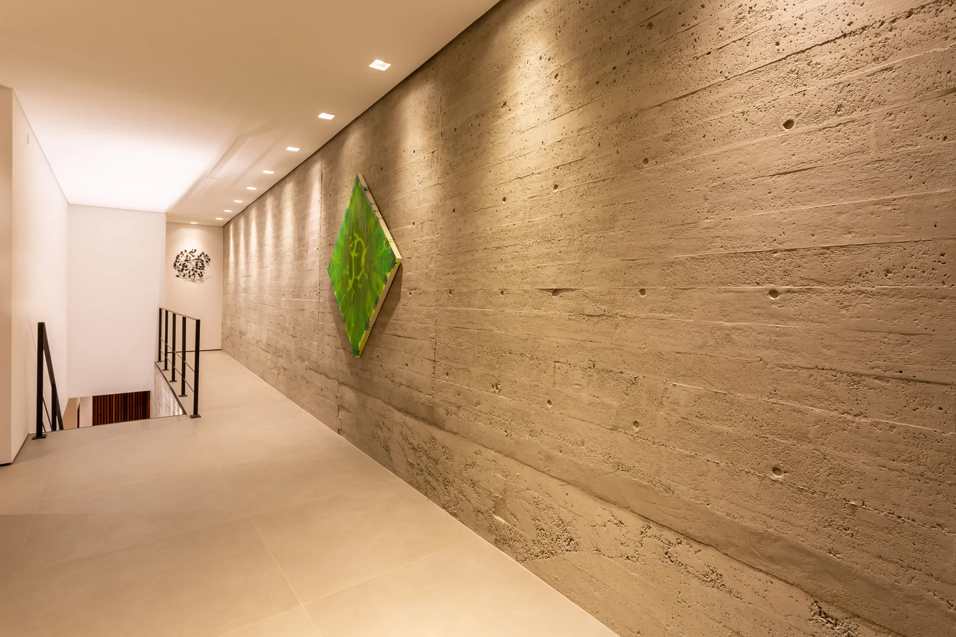 A minimalist hallway featuring a long board-formed concrete wall illuminated by recessed ceiling spotlights, with a green diamond-shaped art piece.