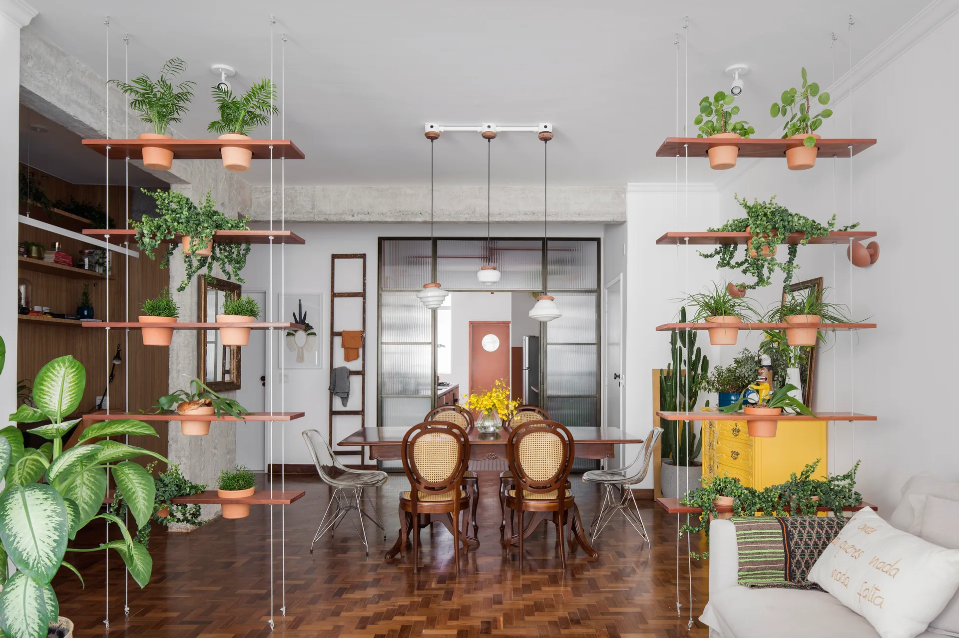 A dining room features a wooden table and cane-back chairs positioned between two suspended shelving units holding numerous potted plants.
