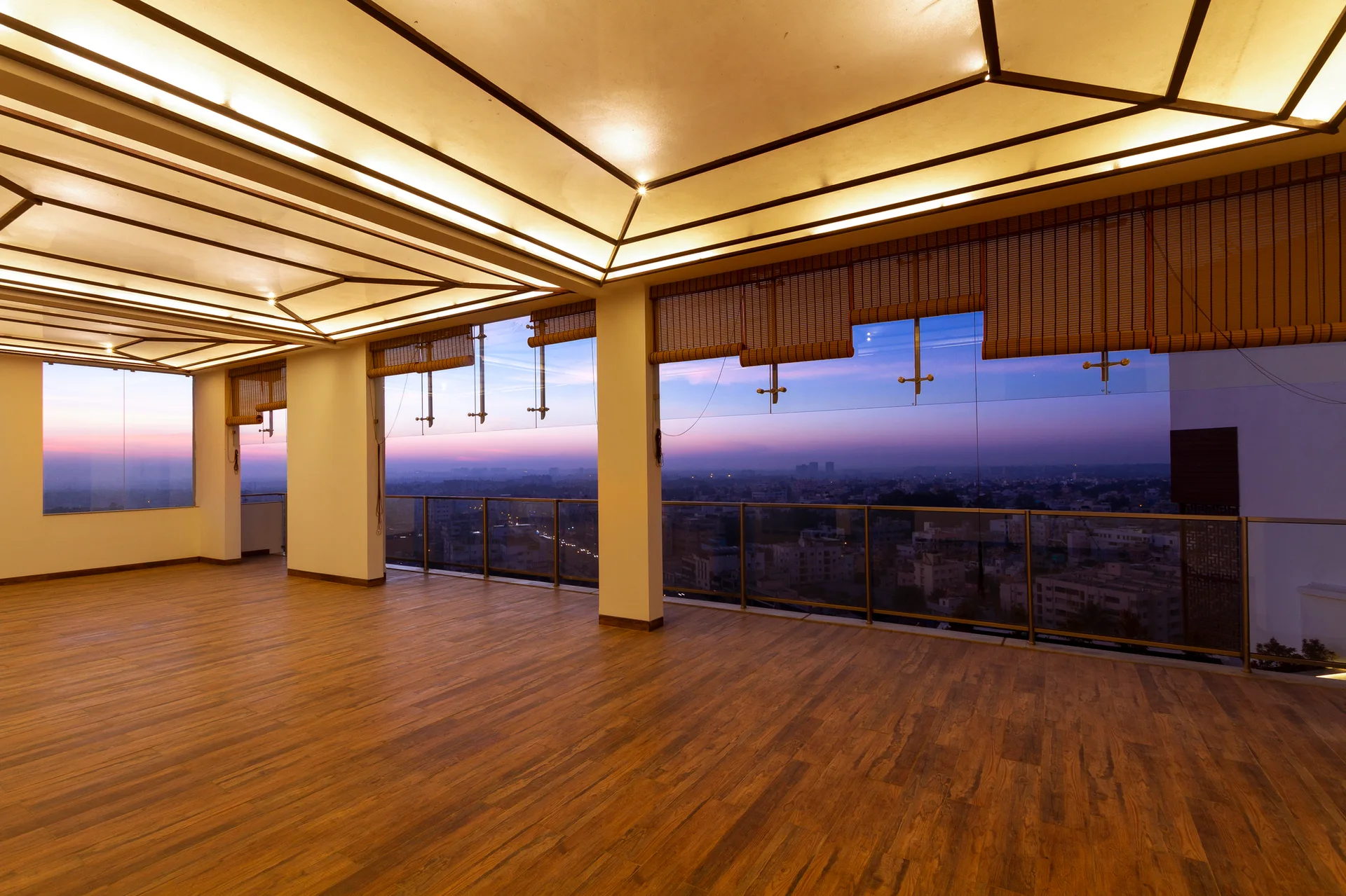 An empty room features wide wood plank flooring, a geometric lit grid ceiling, and floor-to-ceiling glass windows fitted with rolled bamboo shades.