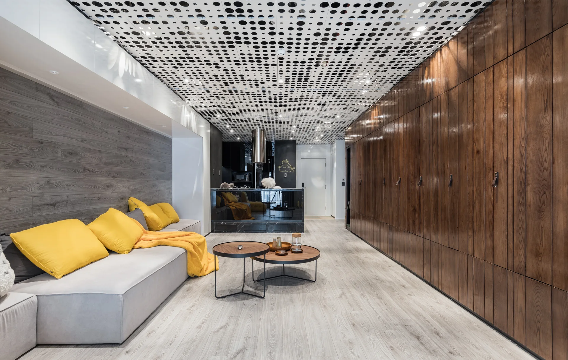 A modern living room features a grey sectional sofa with yellow pillows, a large wood storage cabinet wall, and a patterned perforated ceiling.
