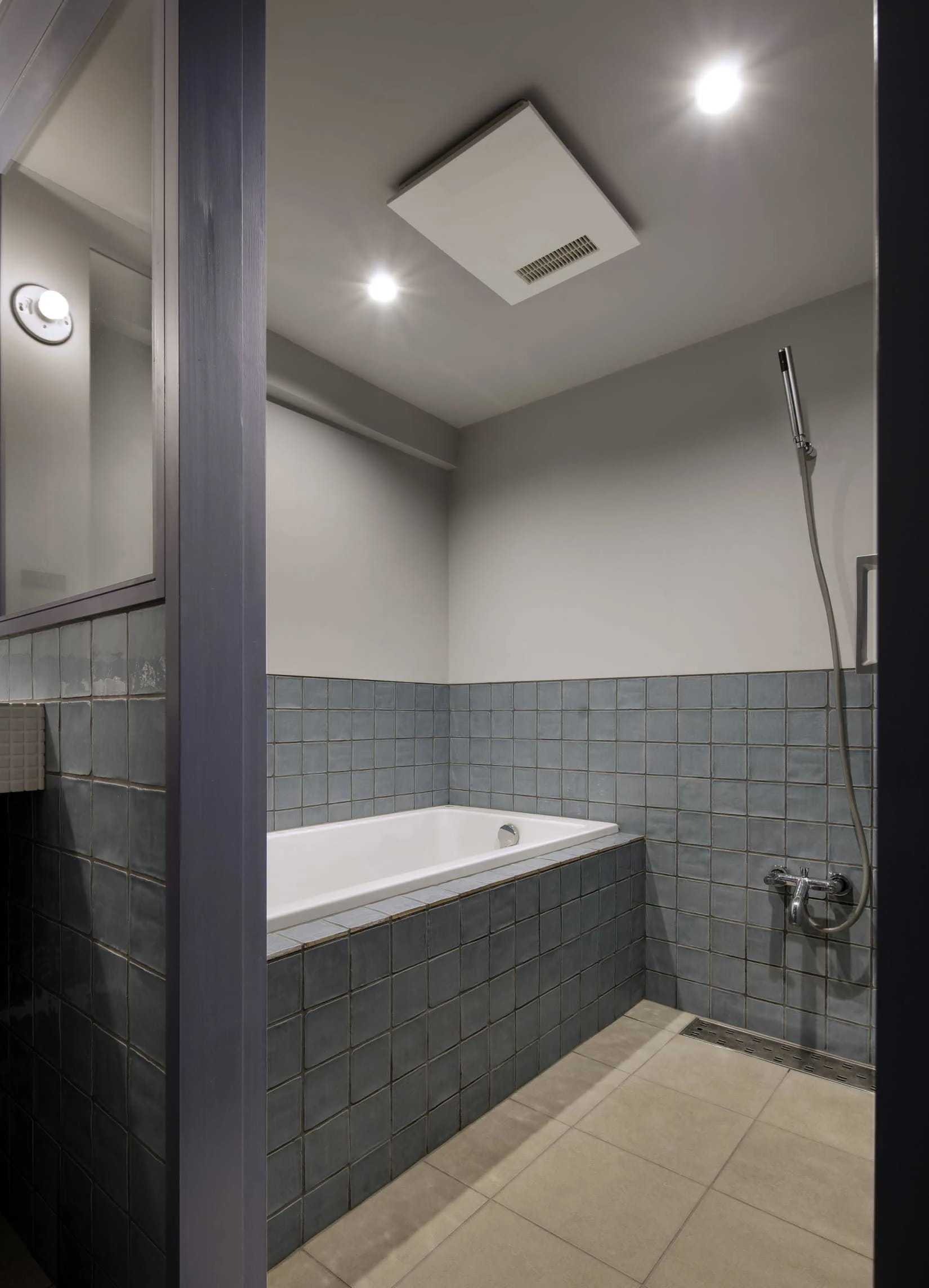 A contemporary bathroom features a built-in white bathtub surrounded by blue-grey grid tiles and a chrome handheld shower attachment.