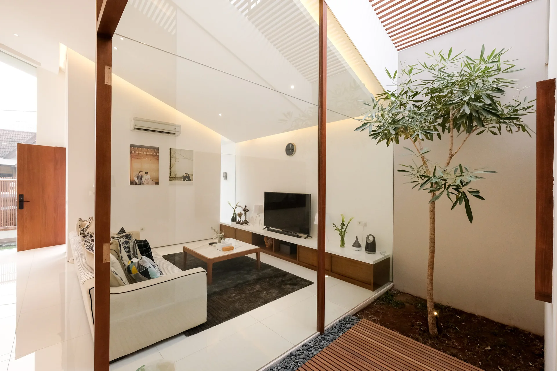 A contemporary living room featuring a cream sofa and low media console is separated from an indoor courtyard by a large glass partition.