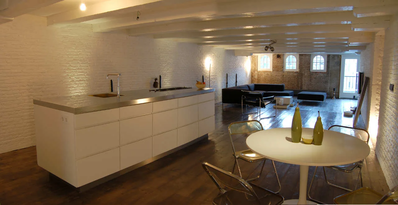 An open loft features a white kitchen island, a round dining table with transparent chairs, and white painted brick walls with exposed beams.