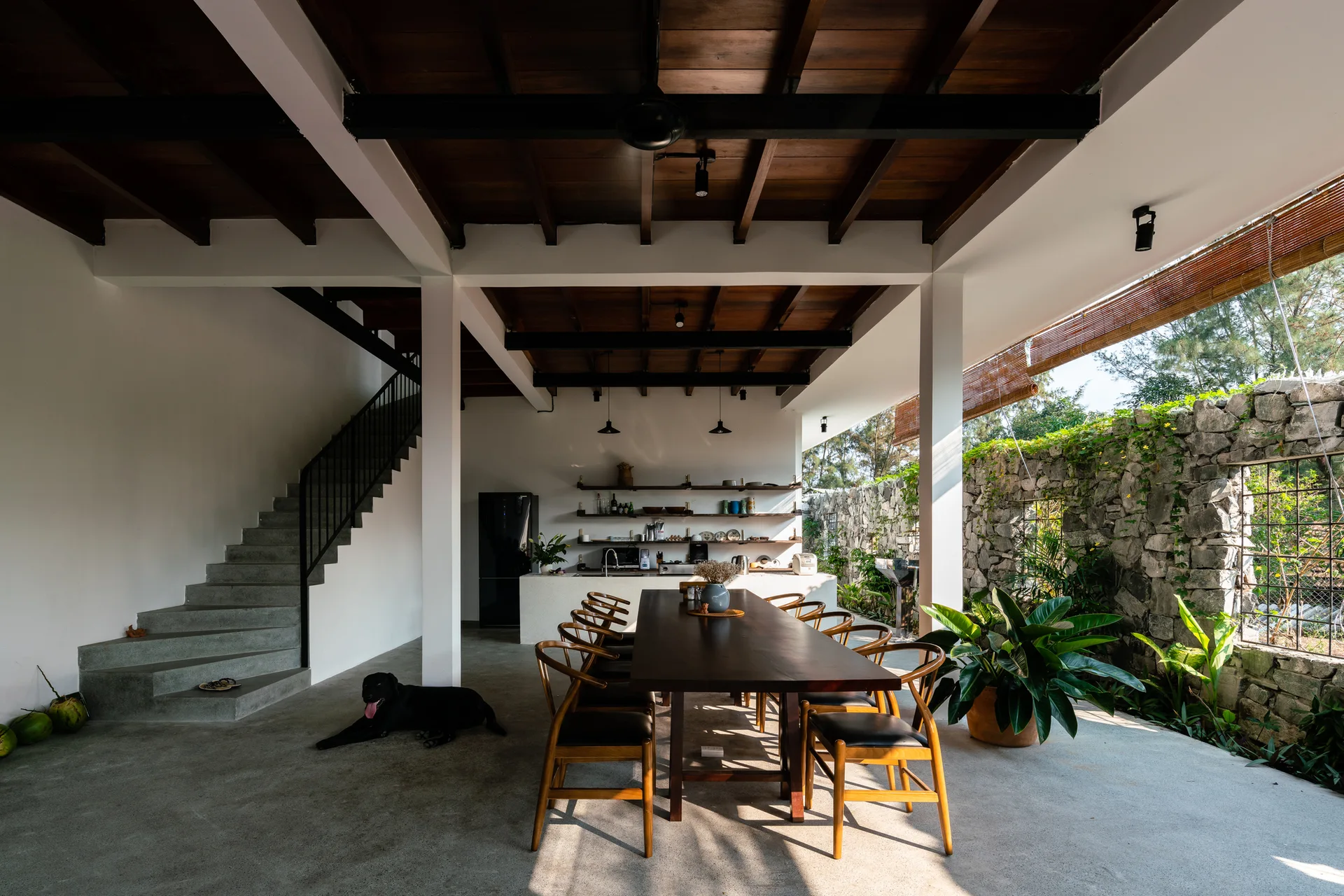 An open-concept dining room with a long wood table, curved chairs, a concrete staircase, and an adjacent stone garden wall under exposed beams.