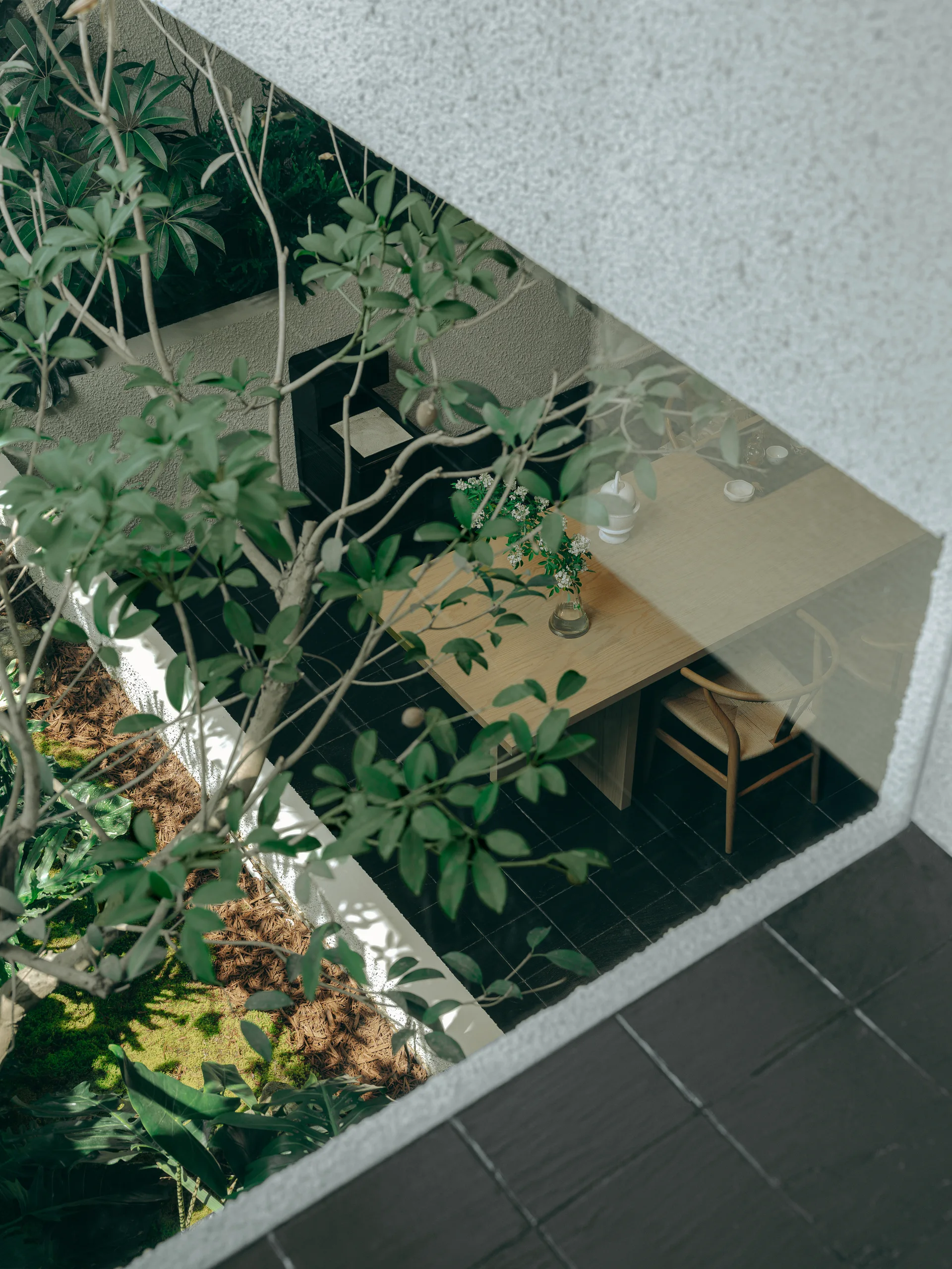 Overhead view of a courtyard dining area with a light wood table, woven-seat chair, and dark tiled floor next to a mossy garden.