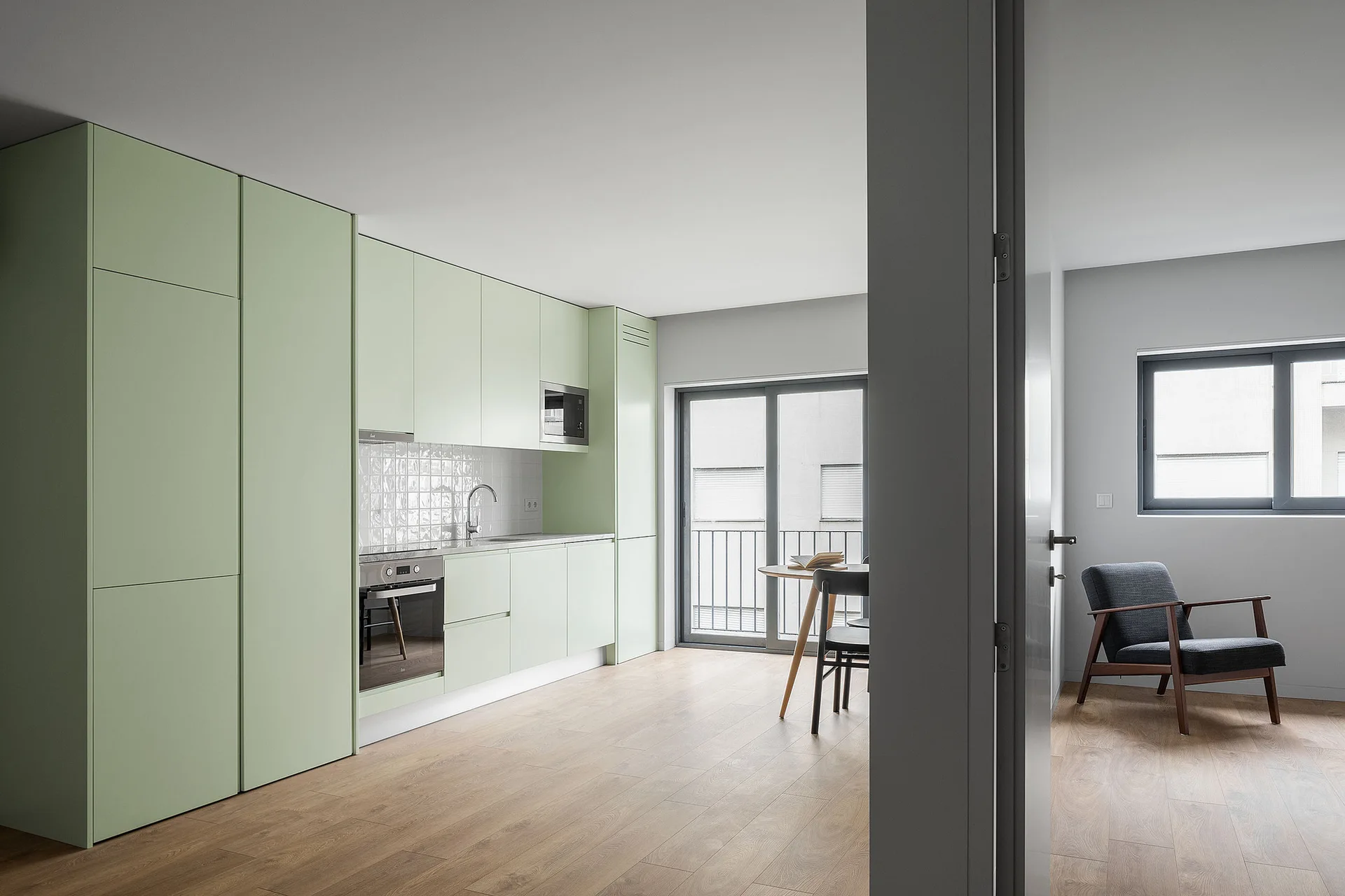 A modern open-plan kitchen with matte pale green handleless cabinets, a white tiled backsplash, wood flooring, and a dining table by a glass door.