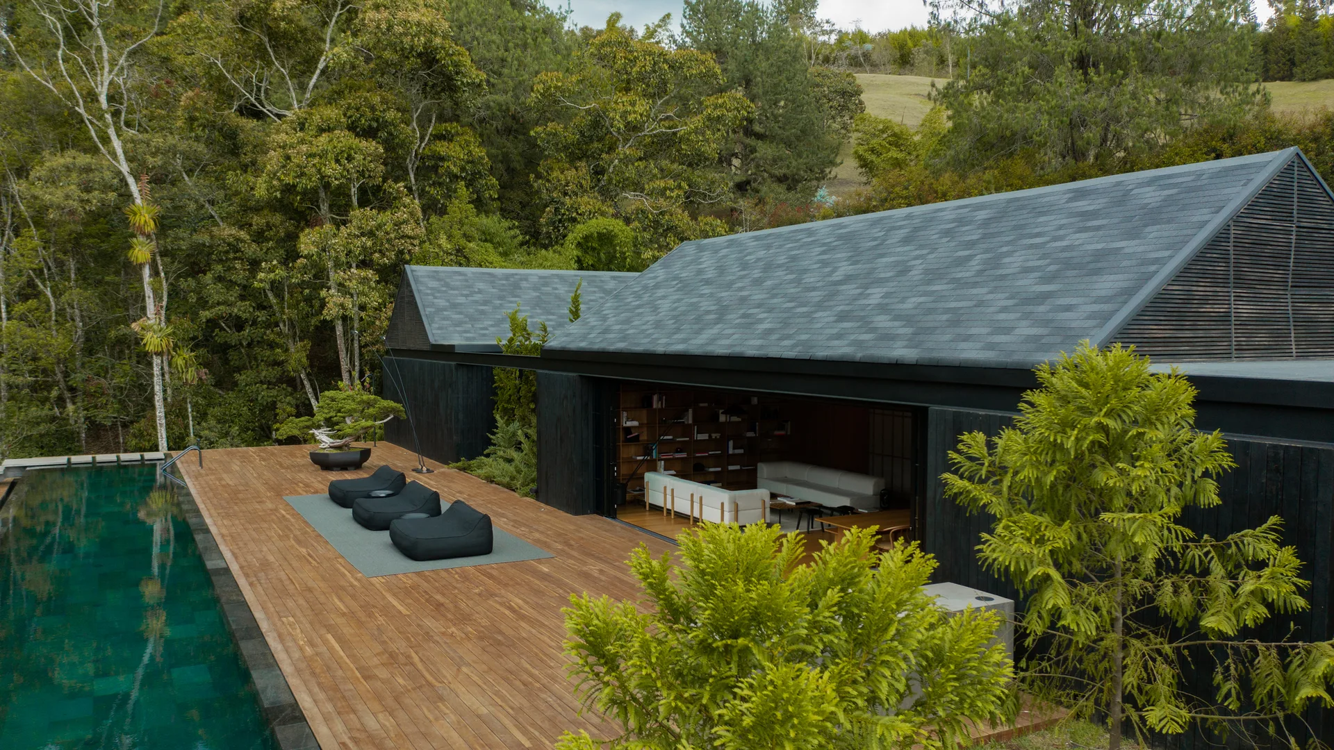 A modern home with black charred wood siding features a wide open living area leading to a wooden deck with black beanbags and an infinity pool.