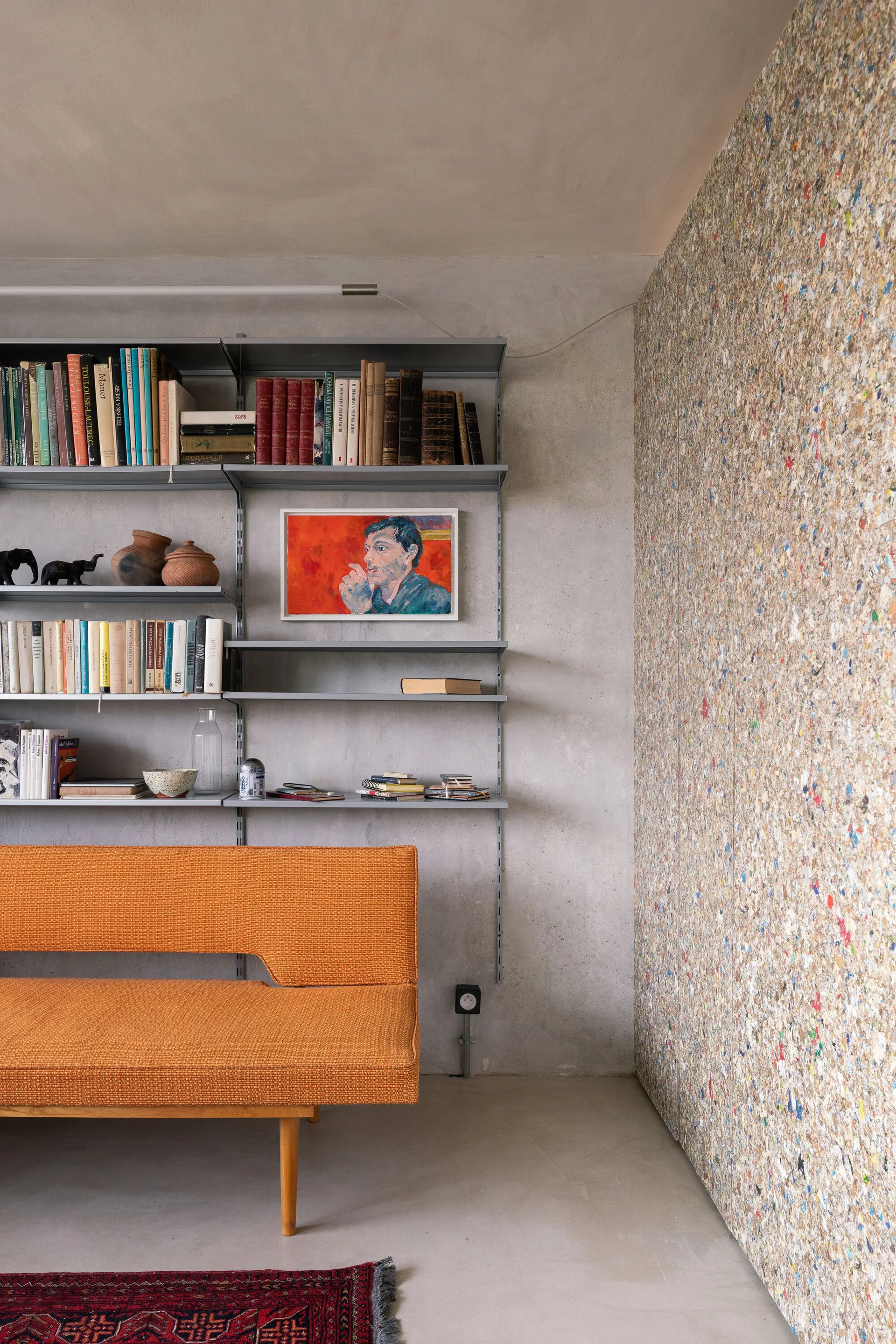 An orange mid-century modern sofa sits in front of a grey metal modular wall shelf filled with books, adjacent to a speckled composite wall panel.