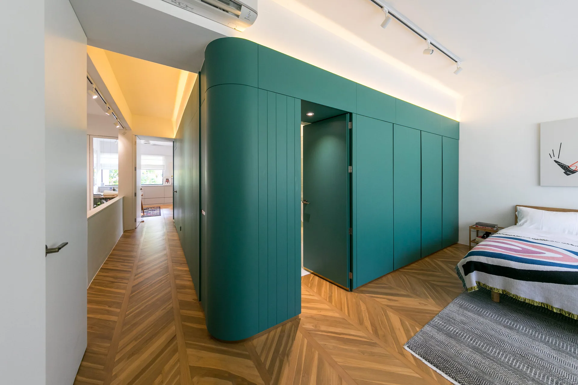 A modern bedroom features a large, curved teal closet wall with an integrated door, set against herringbone wood floors next to a neatly made bed.