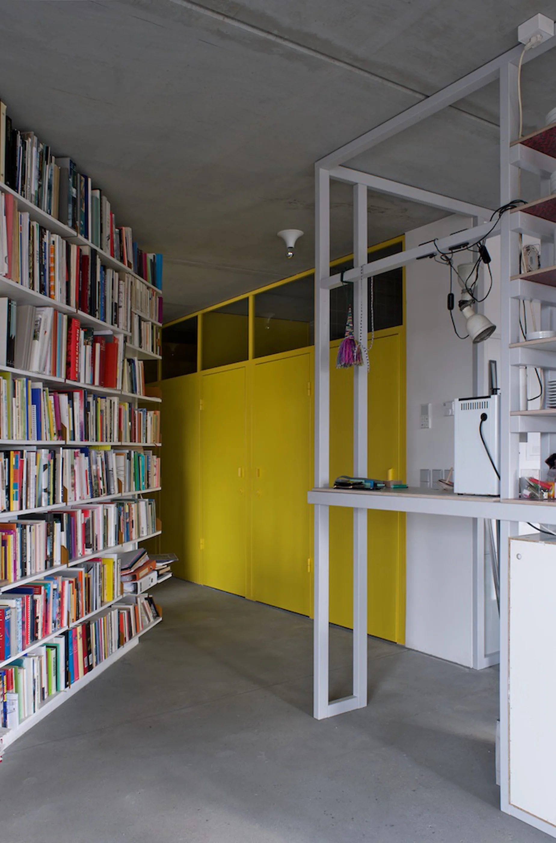 A minimalist home library featuring a tall white bookshelf, an exposed concrete floor, and vibrant yellow storage cabinets in the background.