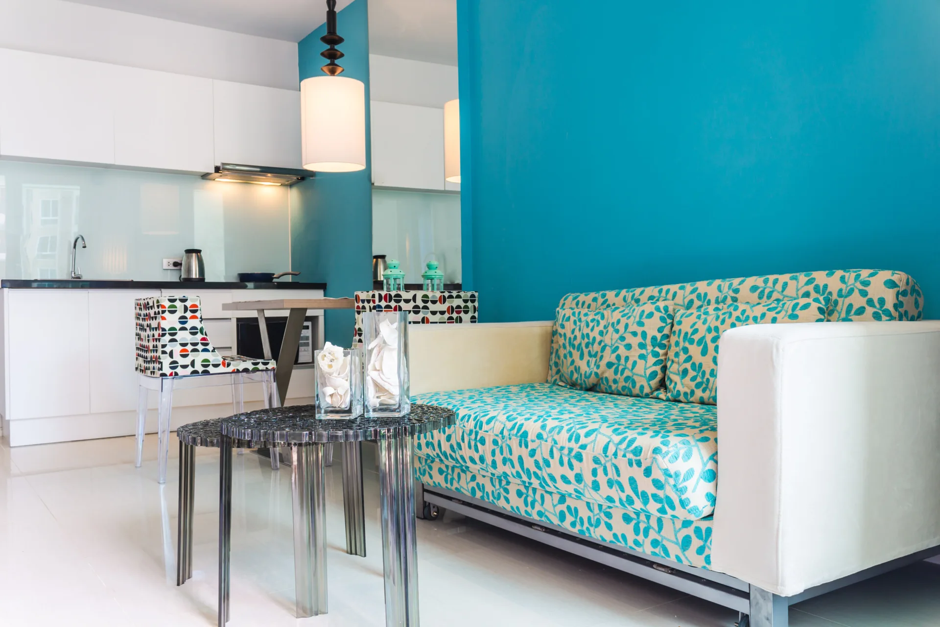 A small living room features a bright blue wall, a patterned turquoise sofa, and clear acrylic tables, with a white kitchen in the background.