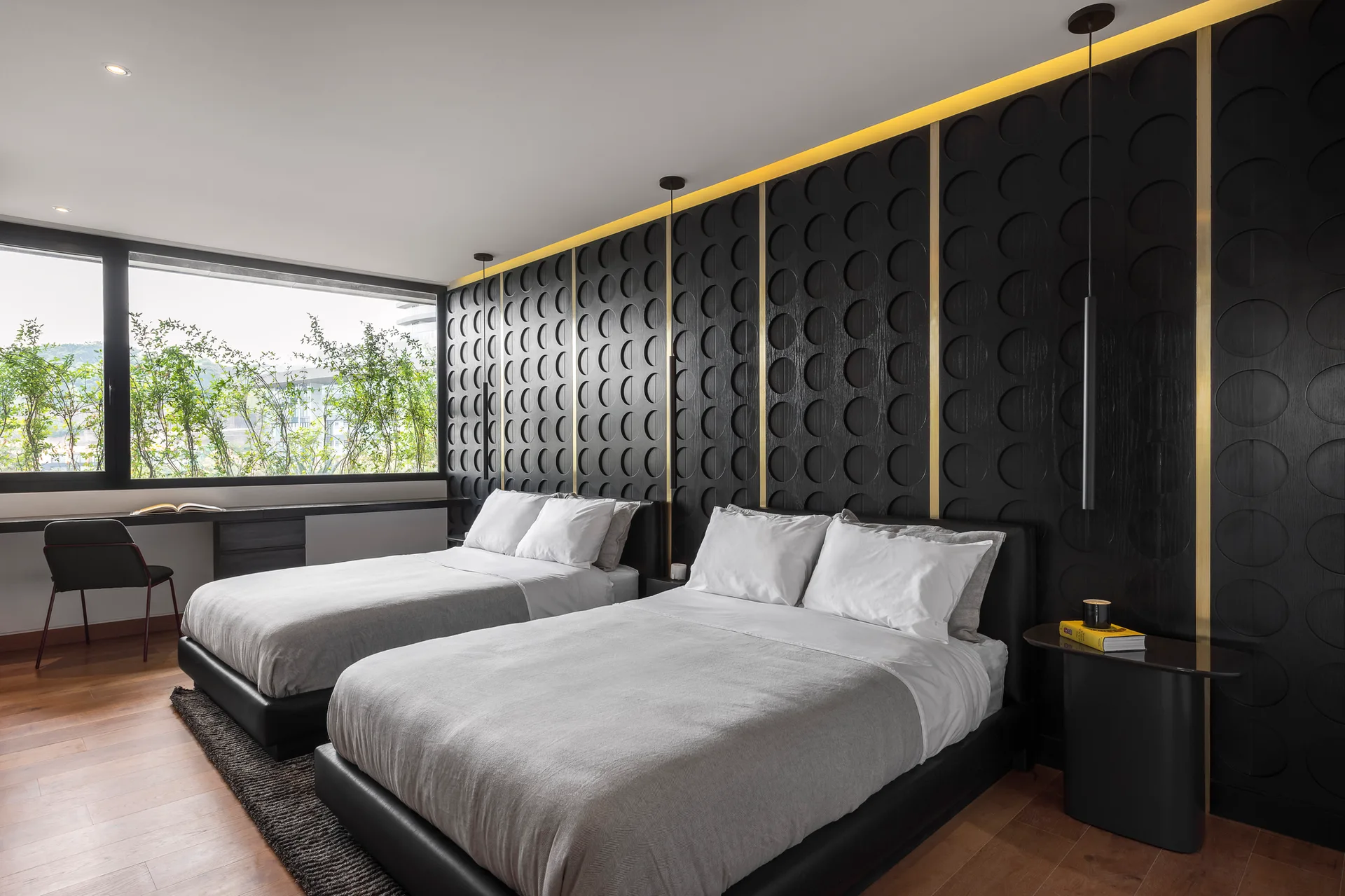 A contemporary bedroom features two beds positioned against a black textured wall panel decorated with gold vertical stripes and a warm glowing light cove.