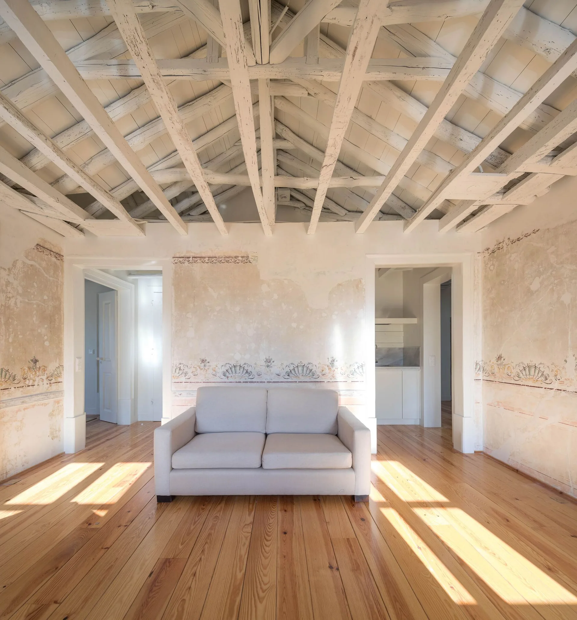 A light grey sofa sits in a rustic room with distressed plaster walls, a faded painted border, and an exposed white-painted wood beamed ceiling.