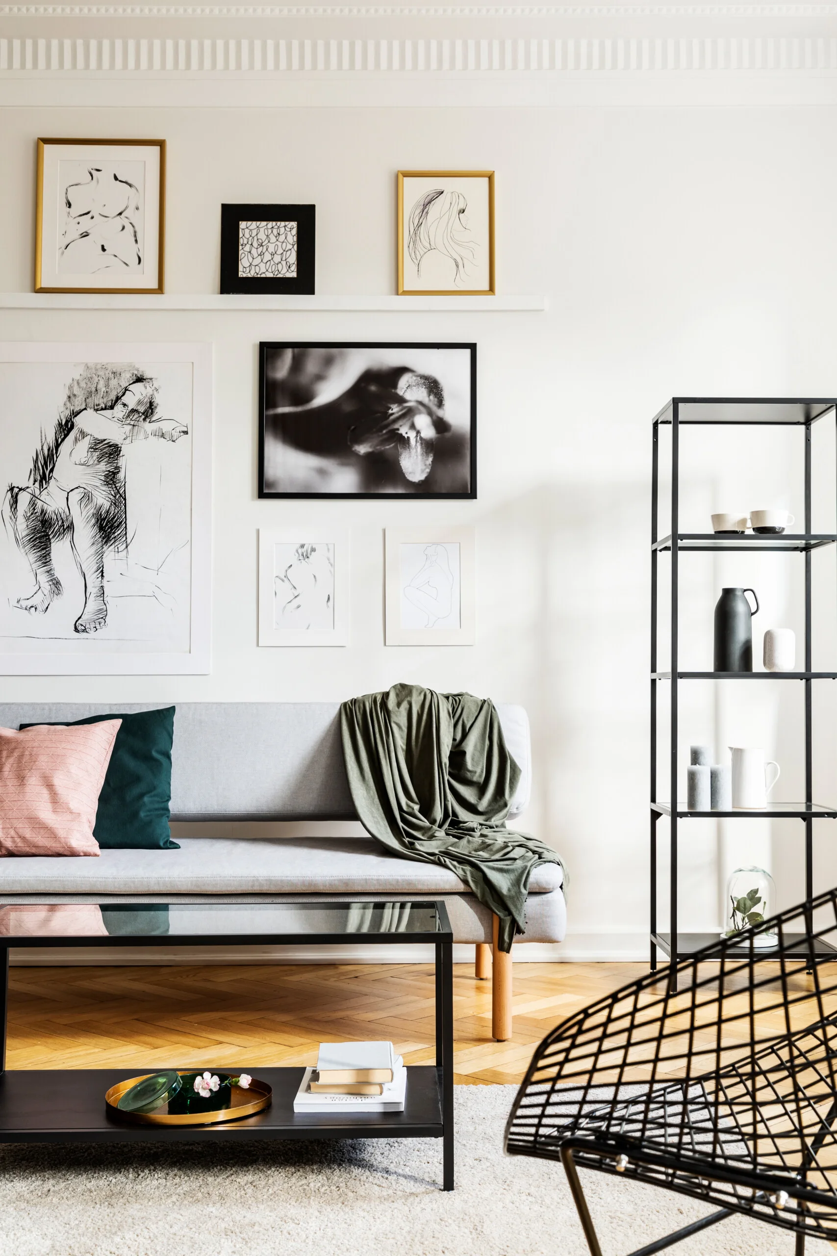 A light grey sofa stands below a gallery wall of framed art prints next to a black metal shelf on herringbone floors.
