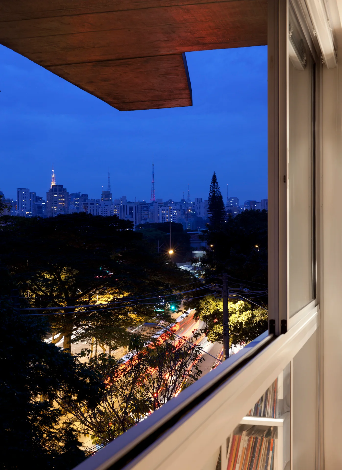 A view through an apartment window looking out over a city at dusk, with books visible in a shelf below the sill.