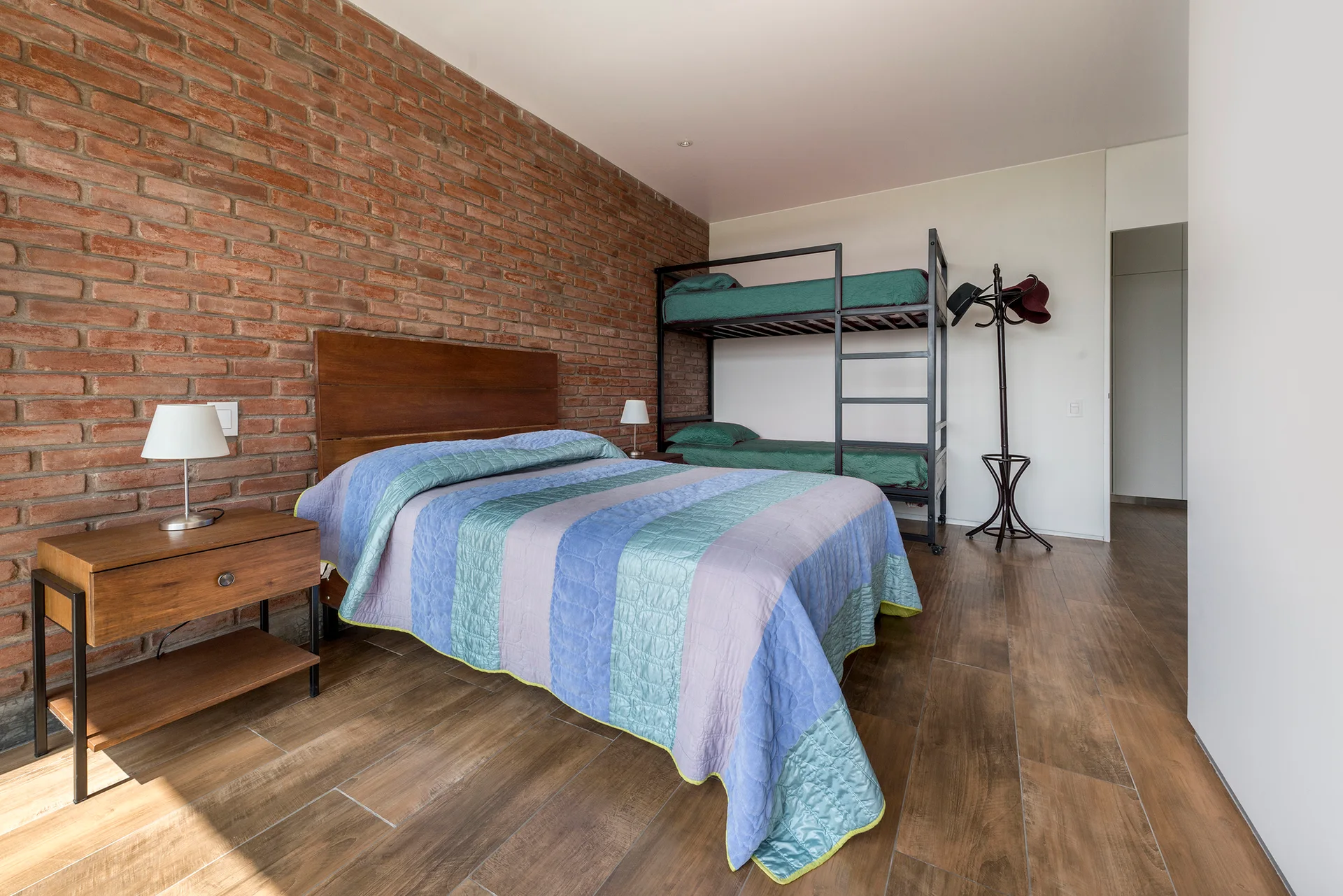 A bedroom features an exposed red brick wall, a wood-framed primary bed, a black metal bunk bed, and wood-look floor tiles.